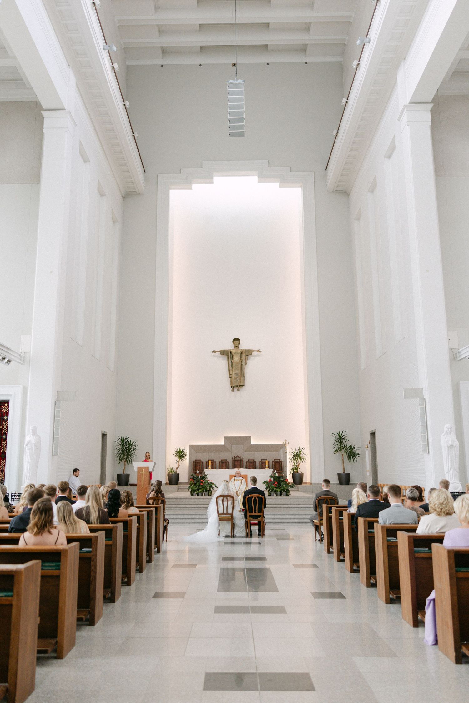 Elegant white church wedding ceremony with guests seated in wooden pews facing the altar with a Christ statue.