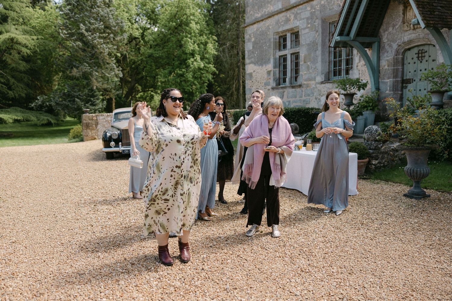 Elegantly dressed women socializing outdoors at a wedding reception near a charming manor house surrounded by lush greenery.