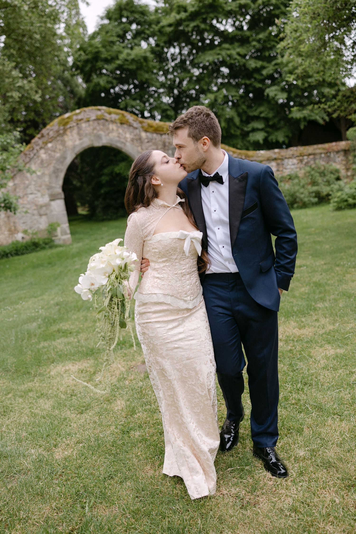 Bride and groom sharing a kiss in garden with stone arch bridge, bride holding white floral bouquet.