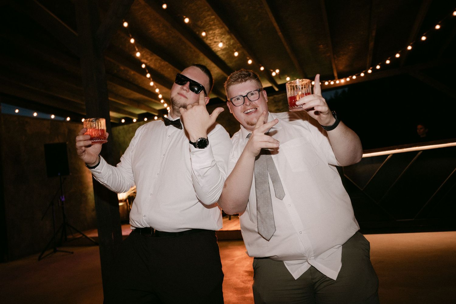 Two groomsmen in white shirts posing with cocktails and sunglasses under string lights at a wedding reception.