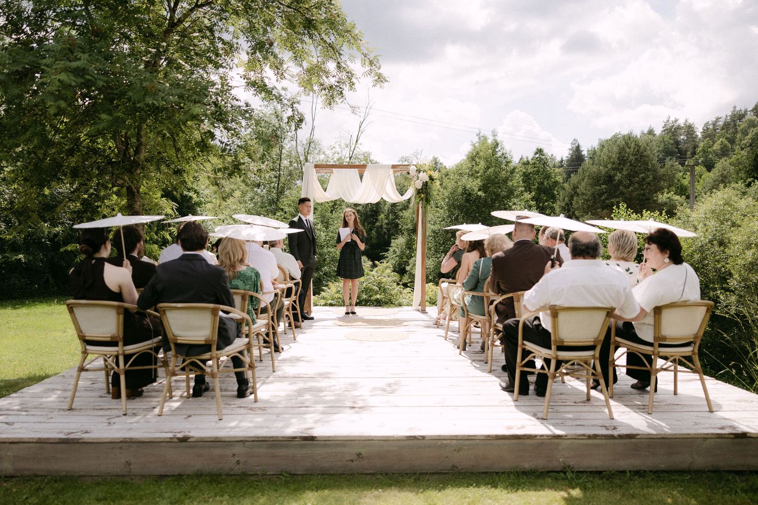Outdoor wedding ceremony on wooden deck with guests seated in rattan chairs under lush green trees.