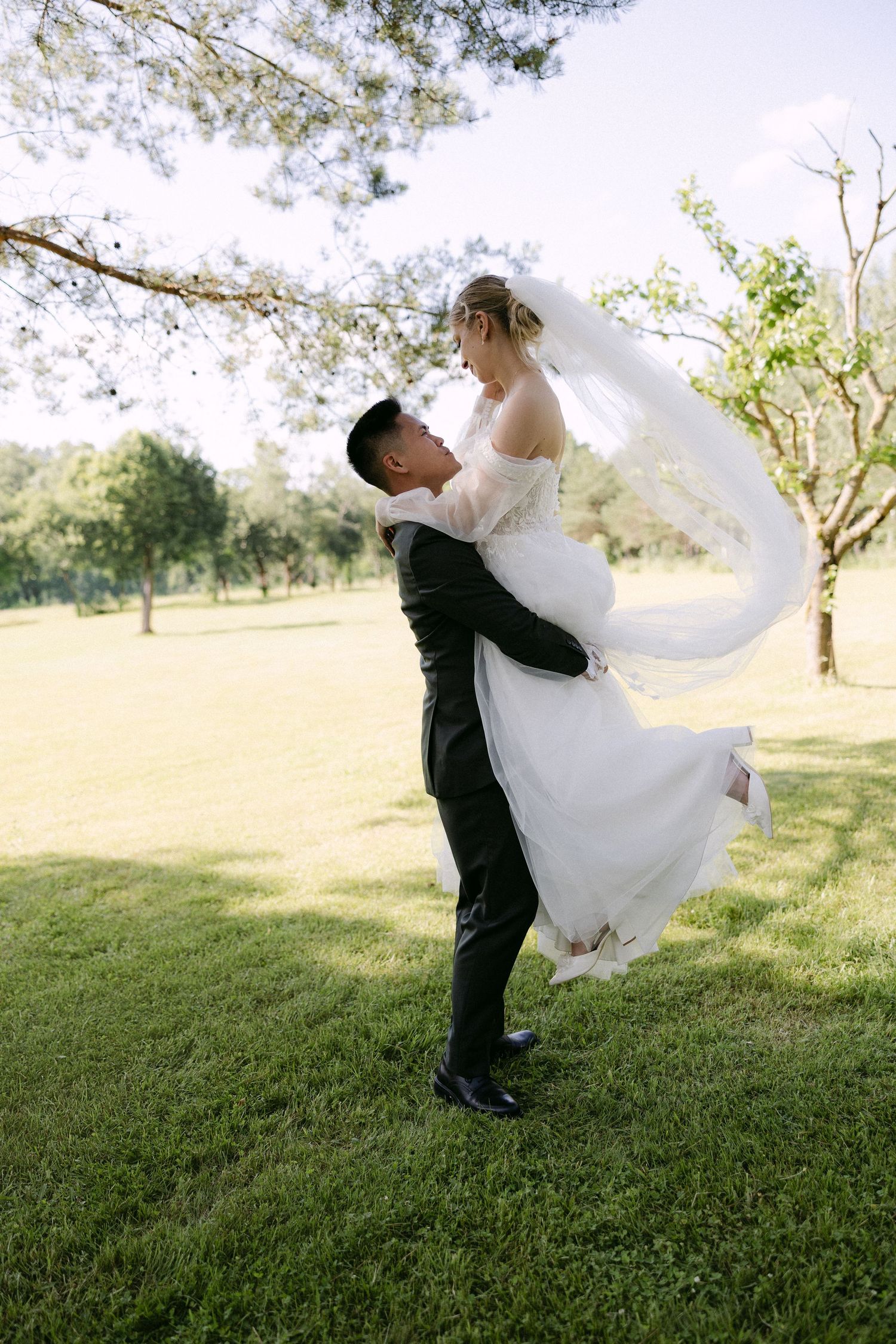 Groom in black tuxedo lifts bride in white wedding dress and veil outdoors on green lawn surrounded by trees.