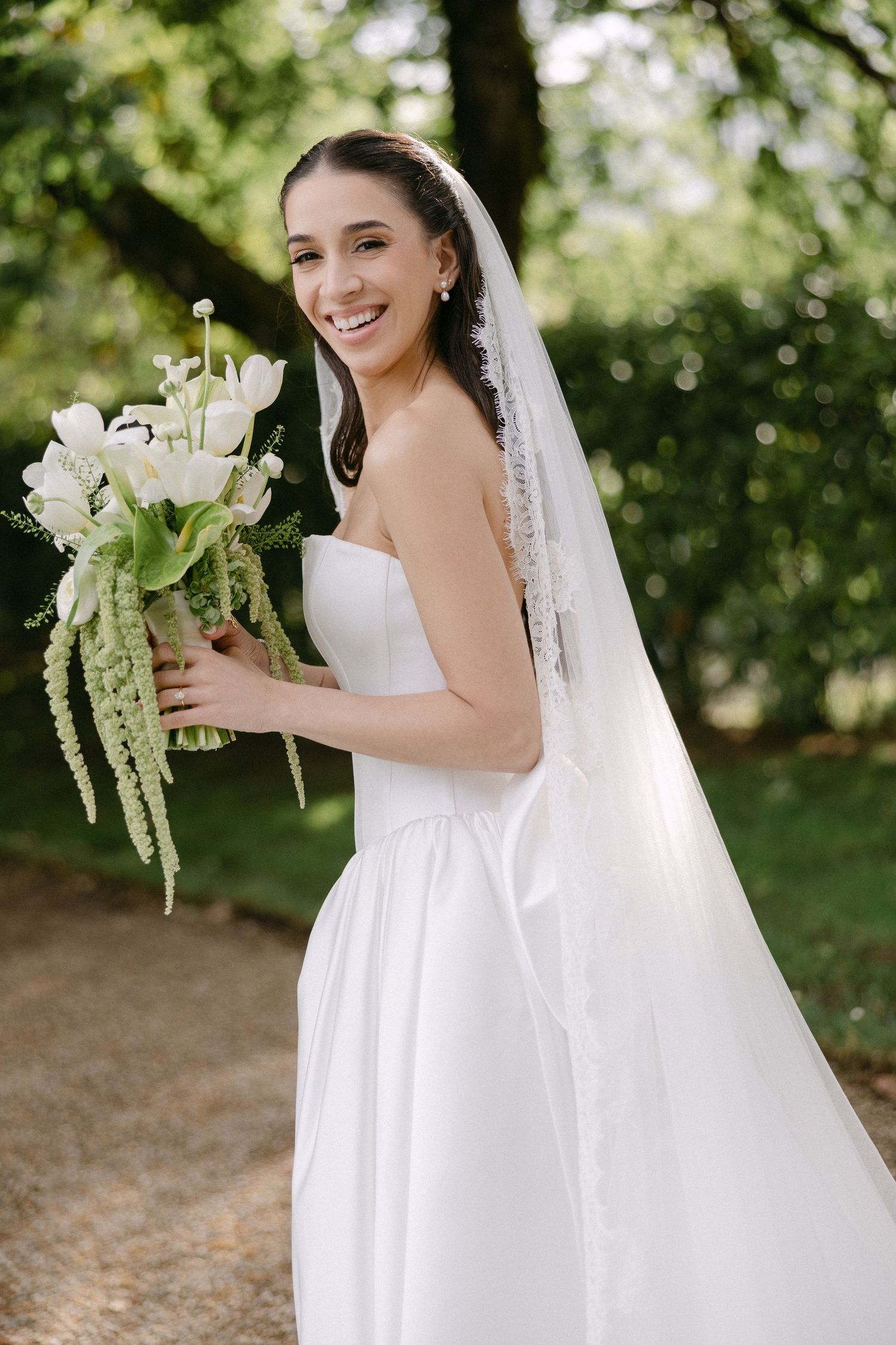 Smiling bride in strapless white gown holds white tulip bouquet with trailing greenery in sunlit garden.