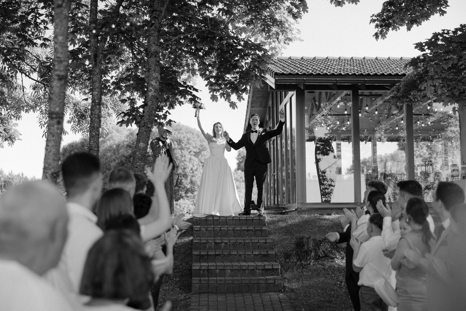 Black and white photo of newlyweds celebrating with raised hands before cheering wedding guests outdoors.