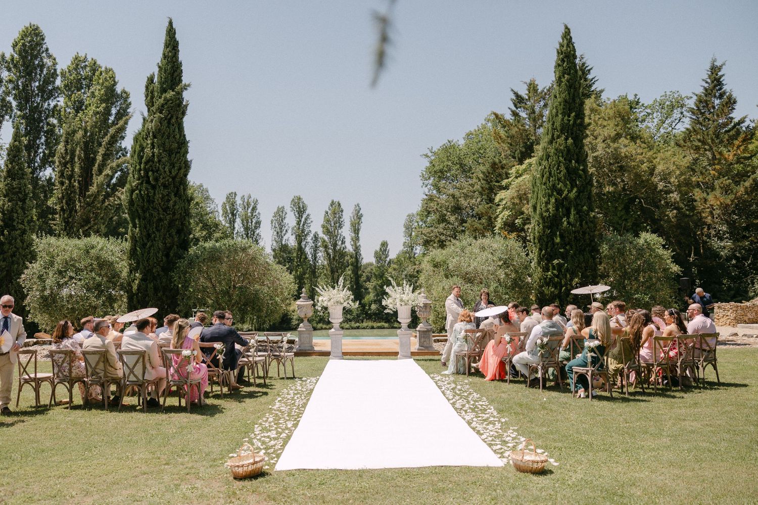 Outdoor wedding ceremony with white aisle runner and guests seated on wooden chairs in manicured garden.