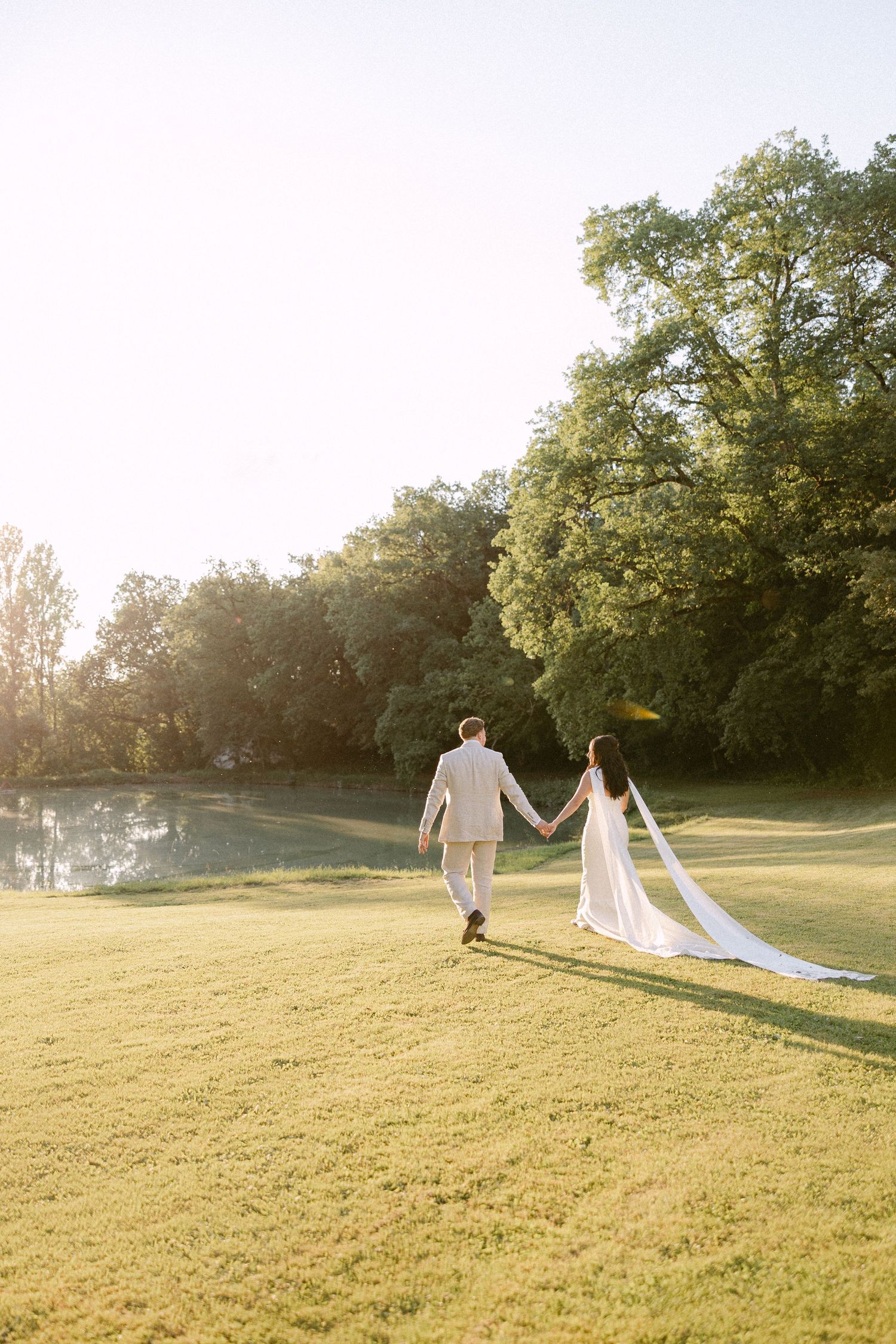 Bride and groom walking hand in hand across sunlit green lawn at golden hour wedding.