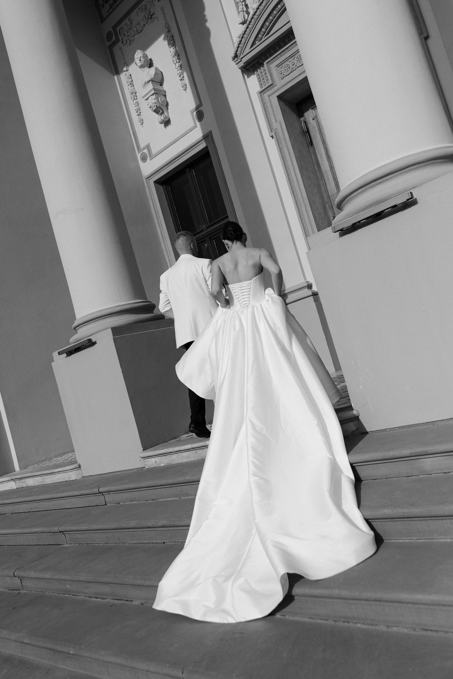 Black and white rear view of bride and groom ascending steps of classical columned building.