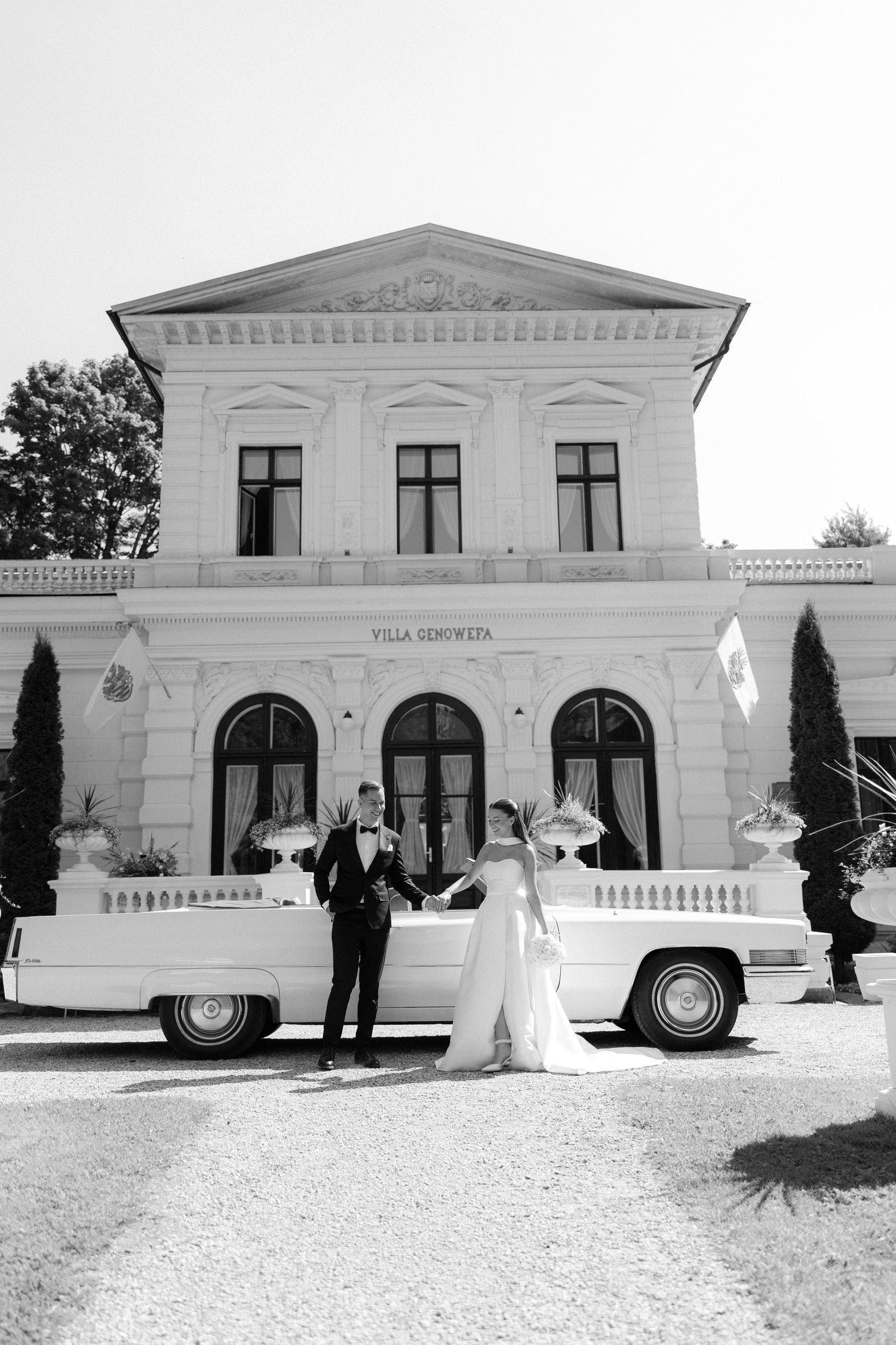 Bride and groom posing by a vintage white car in front of Villa Scenwitta in a classic black and white wedding photo.