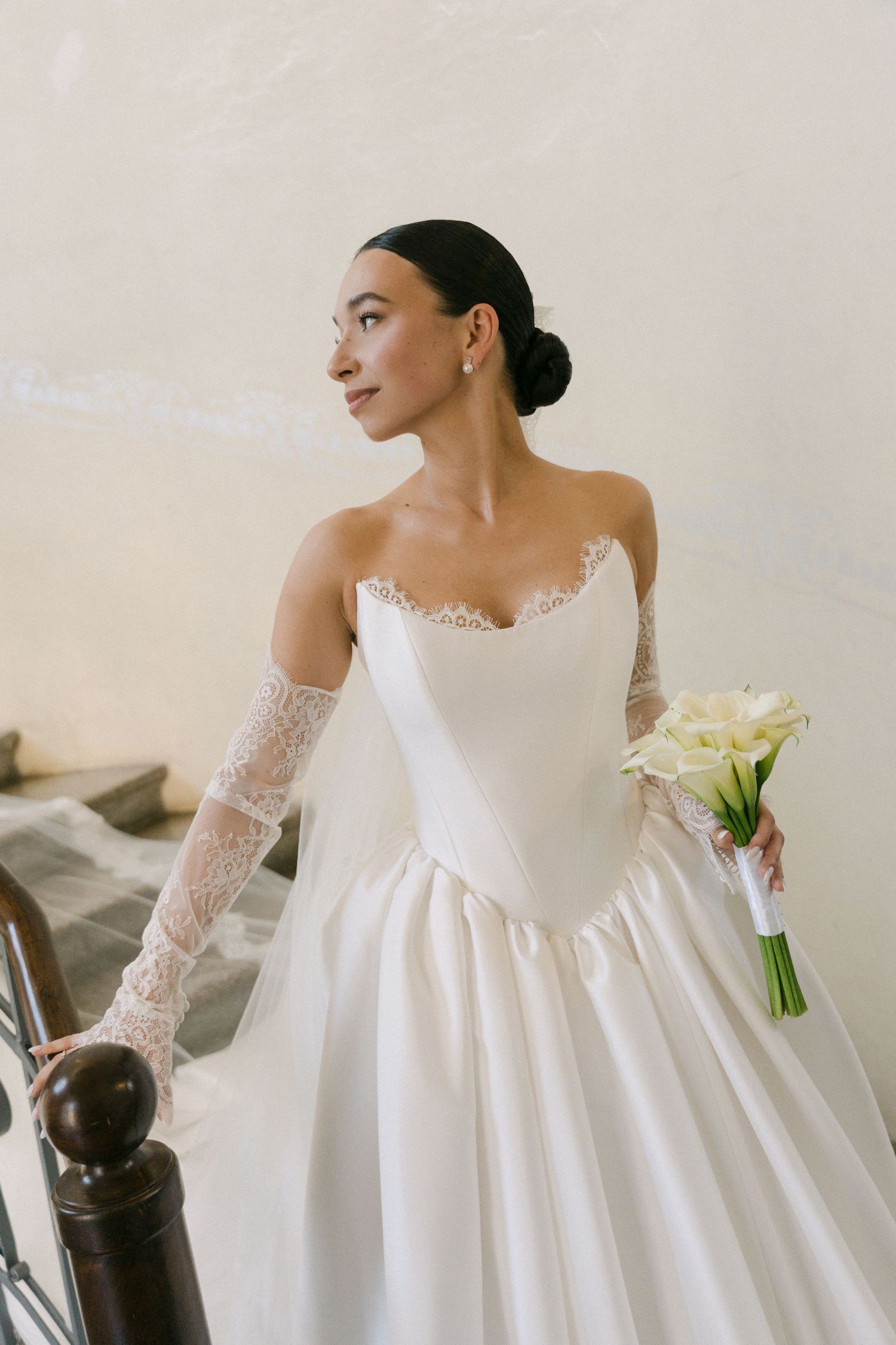 Elegant bride in strapless white ballgown with lace sleeves holding white calla lily bouquet on stairs.
