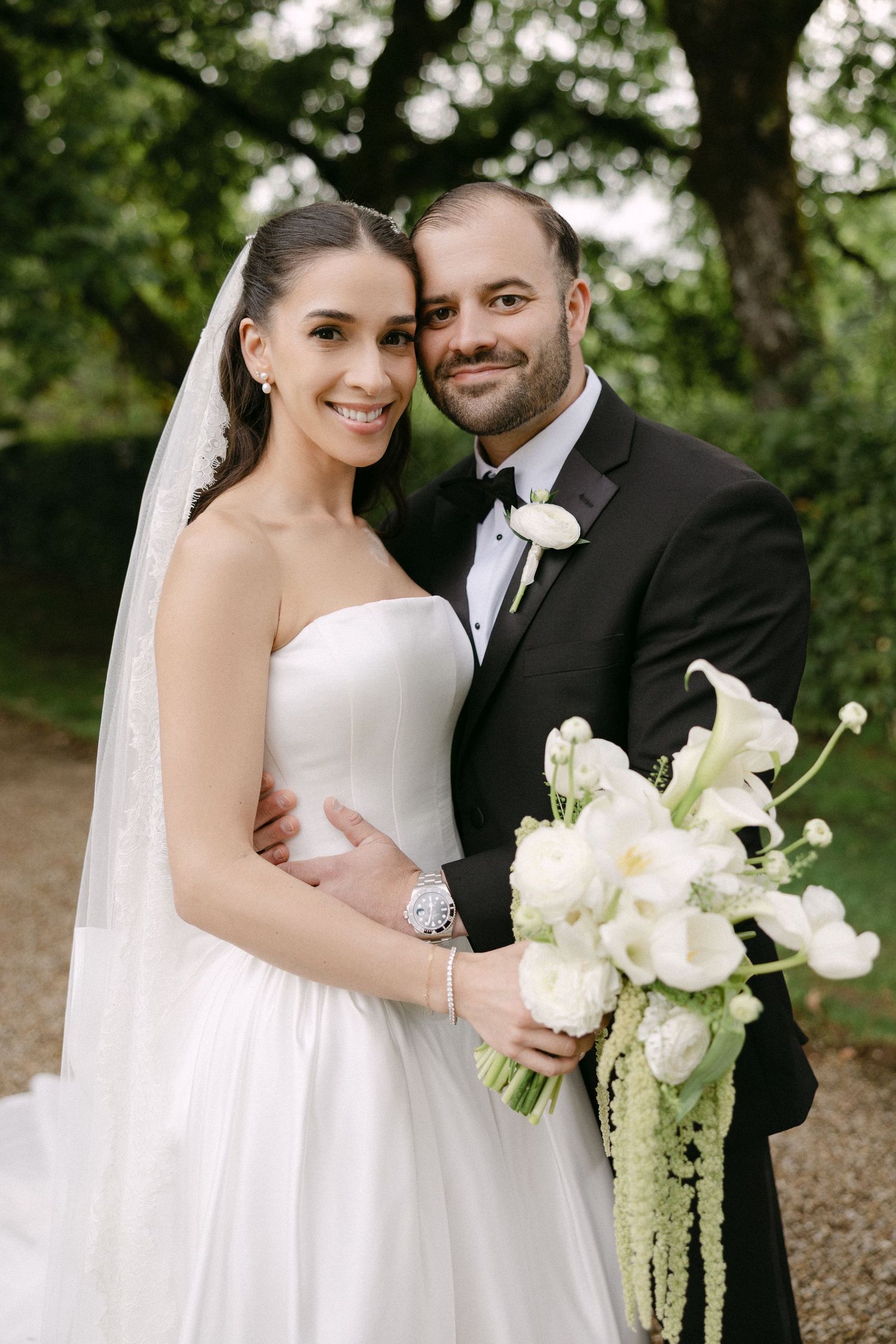 Bride and groom posing outdoors under green trees, bride holding elegant white calla lily bouquet.