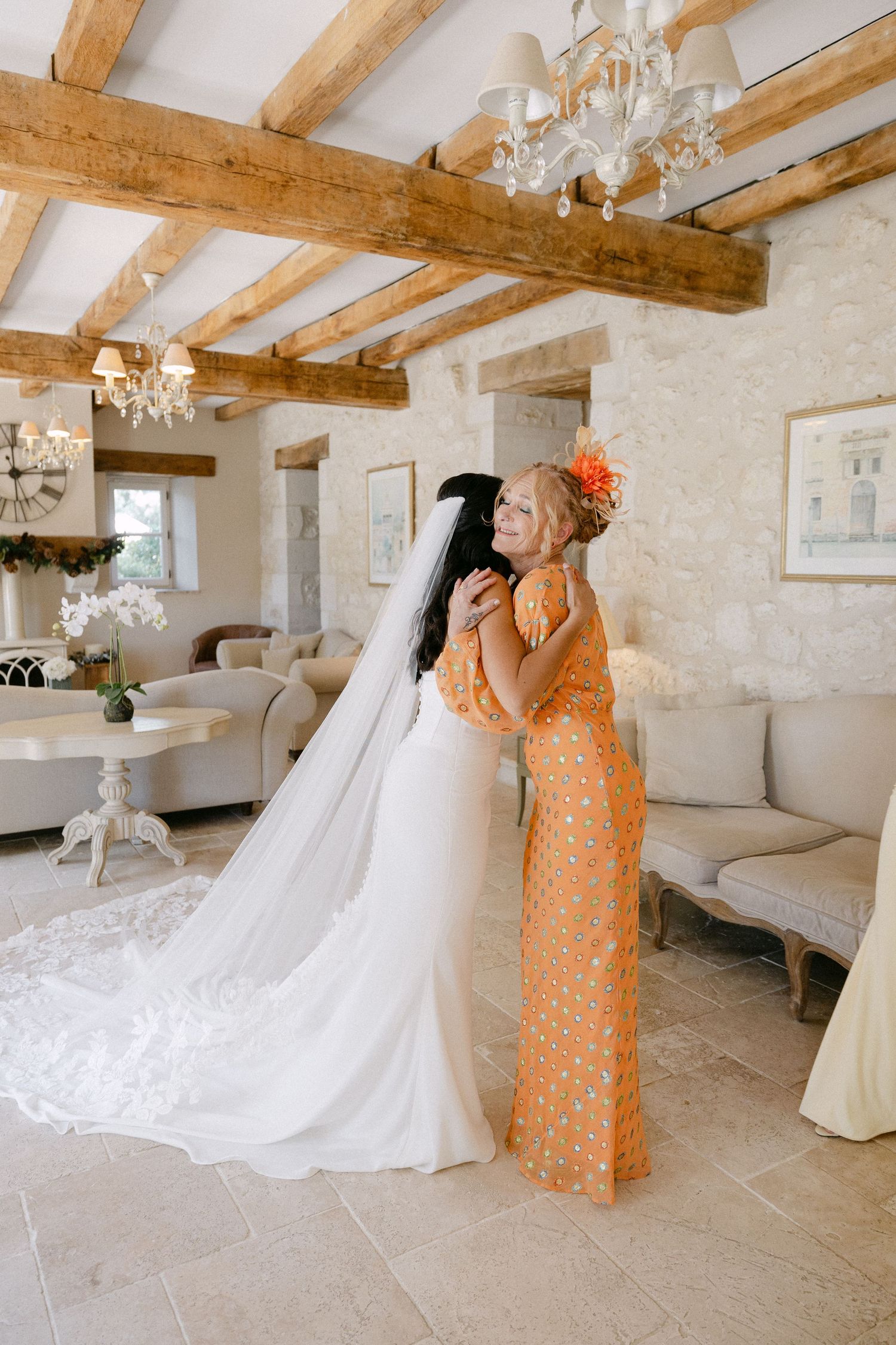 Bride in strapless white gown with veil embracing woman in orange floral dress in French chateau interior.