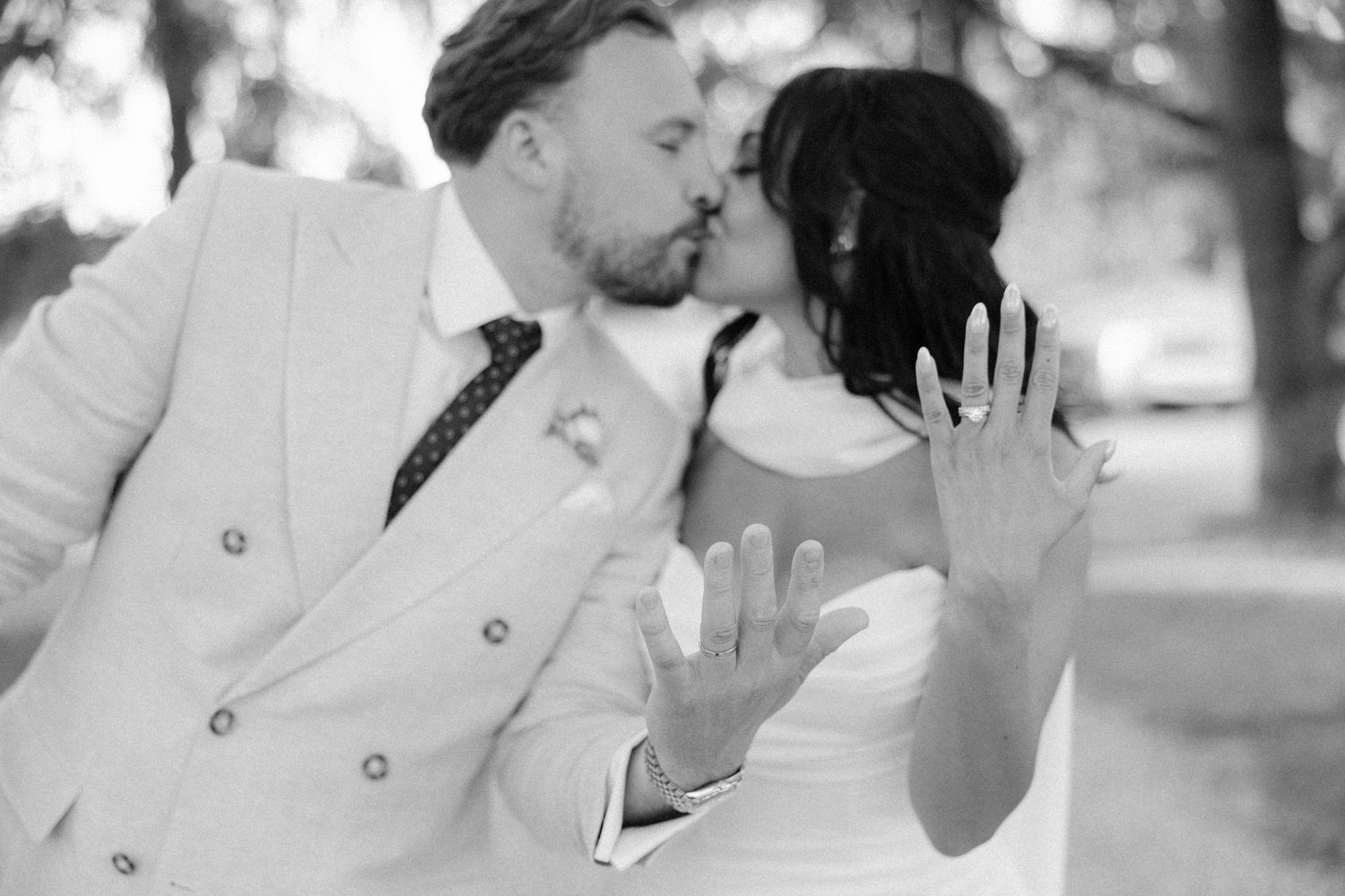Black and white photo of newlyweds kissing and showing wedding rings to camera outdoors.