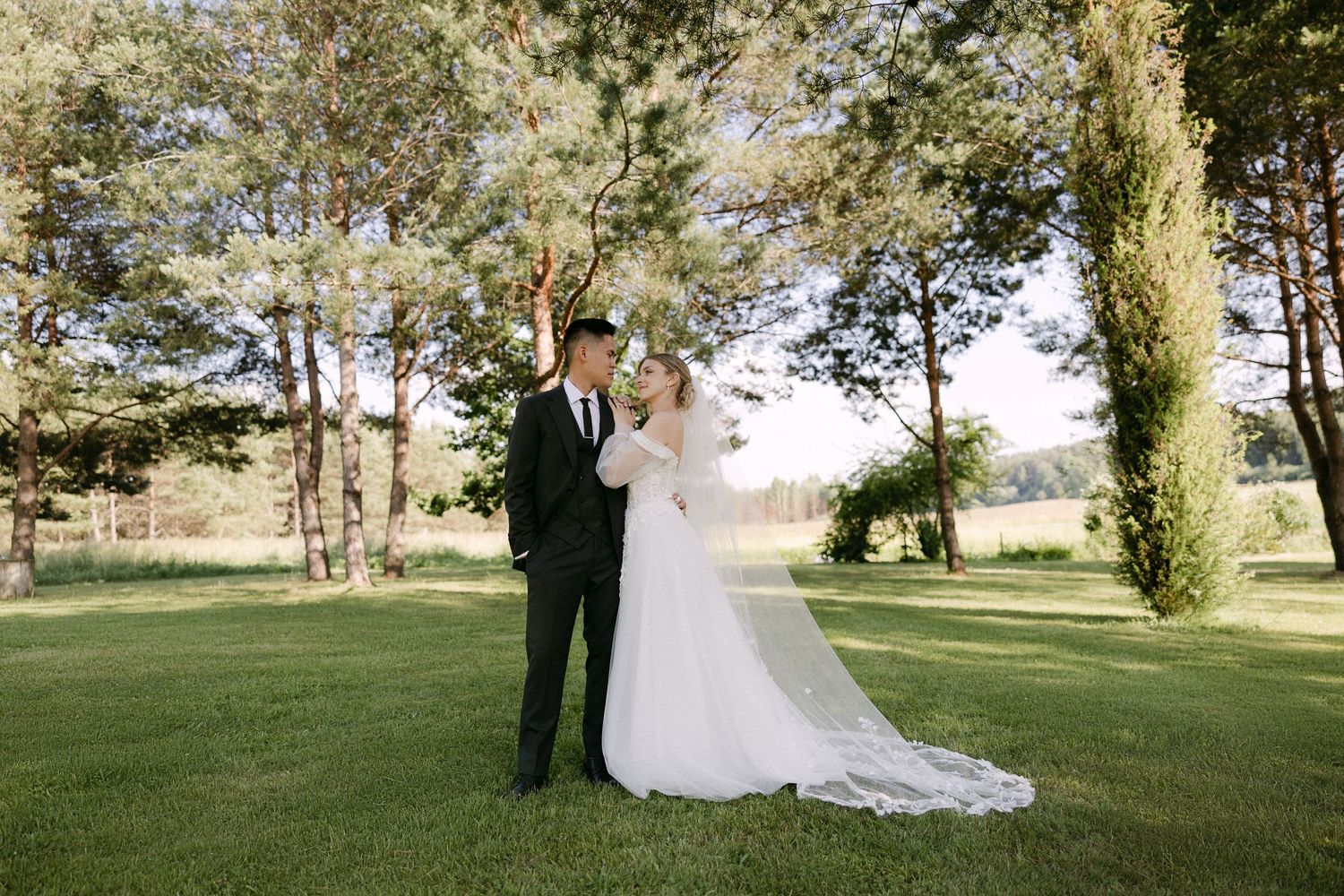 Bride and groom stand together on lush green lawn surrounded by tall trees at outdoor wedding.