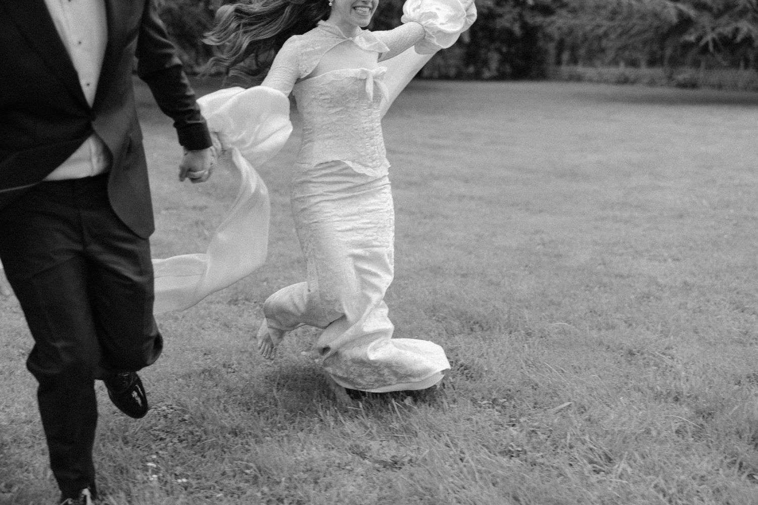 Bride in white gown running across lawn in playful black and white wedding photo.