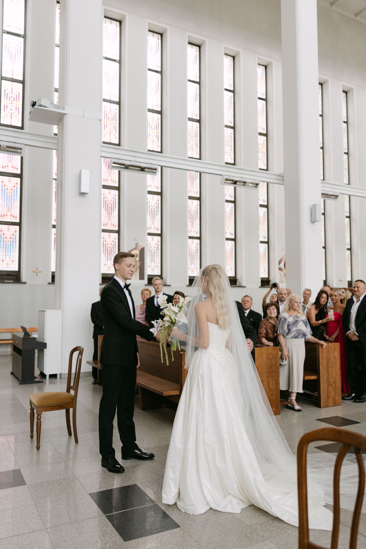 Bride and groom hold hands at the altar during a bright modern church wedding ceremony with guests seated in pews.