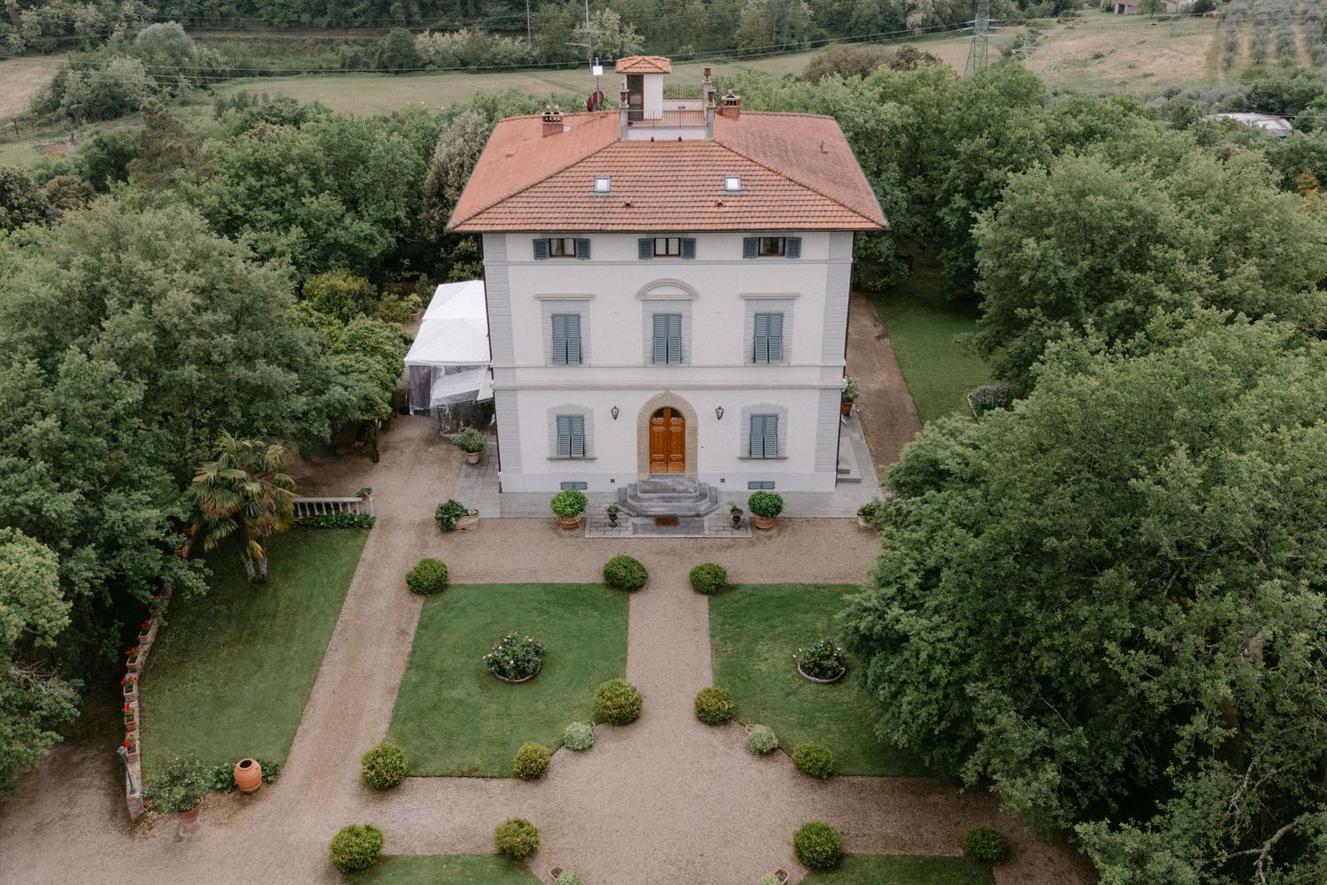 Aerial view of an elegant Italian villa with orange roof, manicured gardens, and surrounding trees.