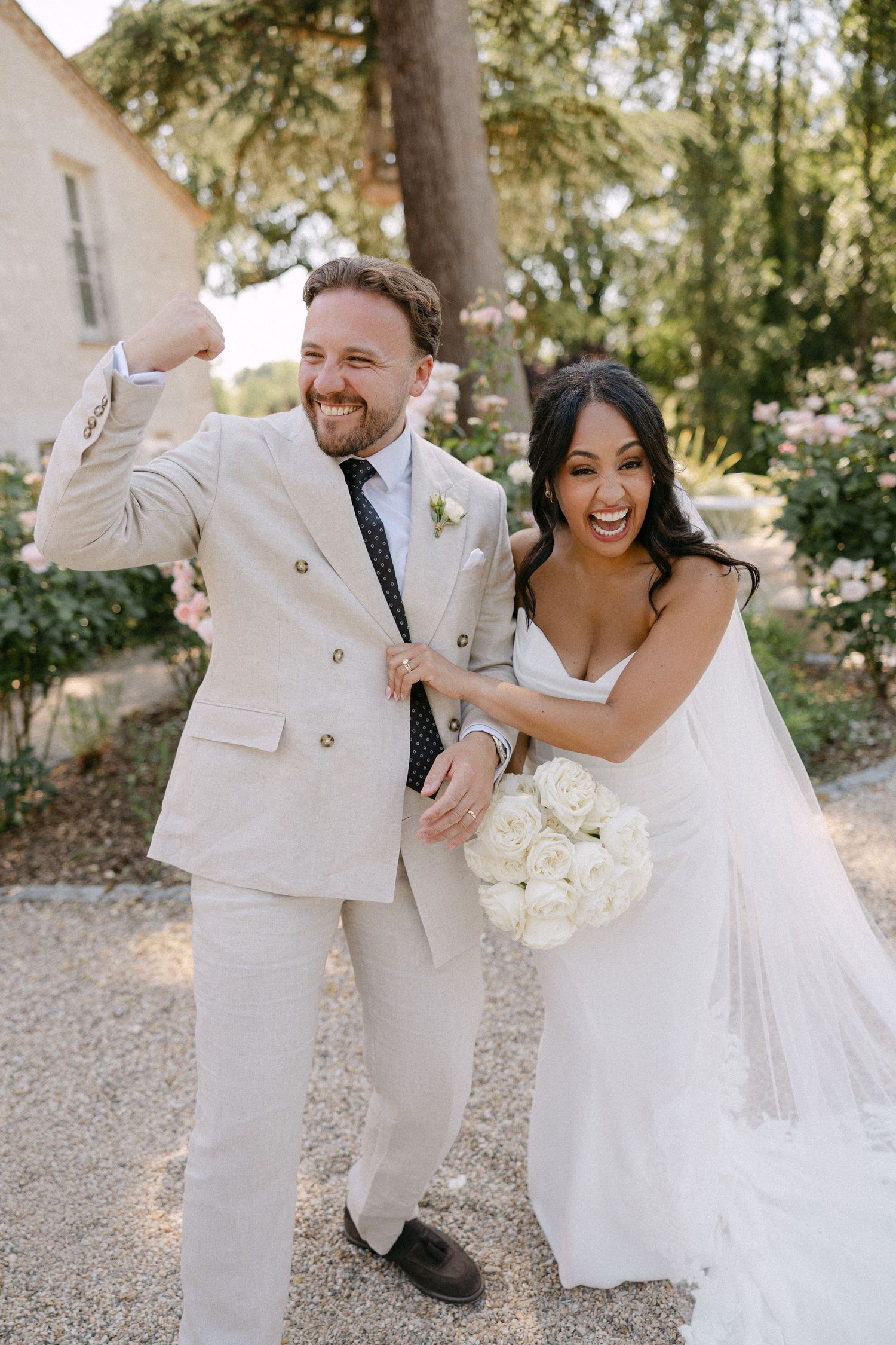 Joyful newlyweds celebrating outdoors, groom in cream suit, bride in white gown holding bouquet.