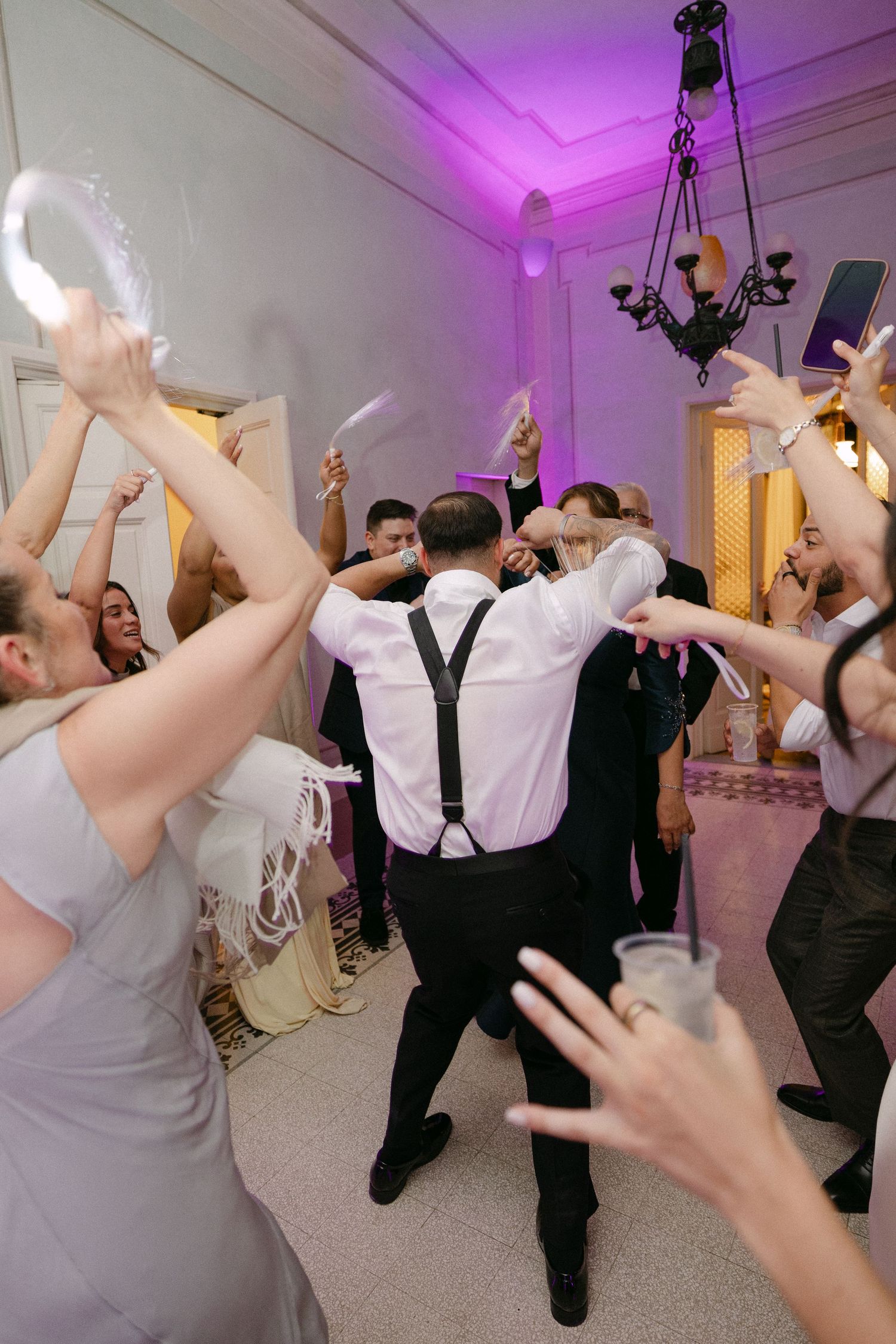 Groom in white shirt and suspenders celebrates with cheering wedding guests on the dance floor at reception.