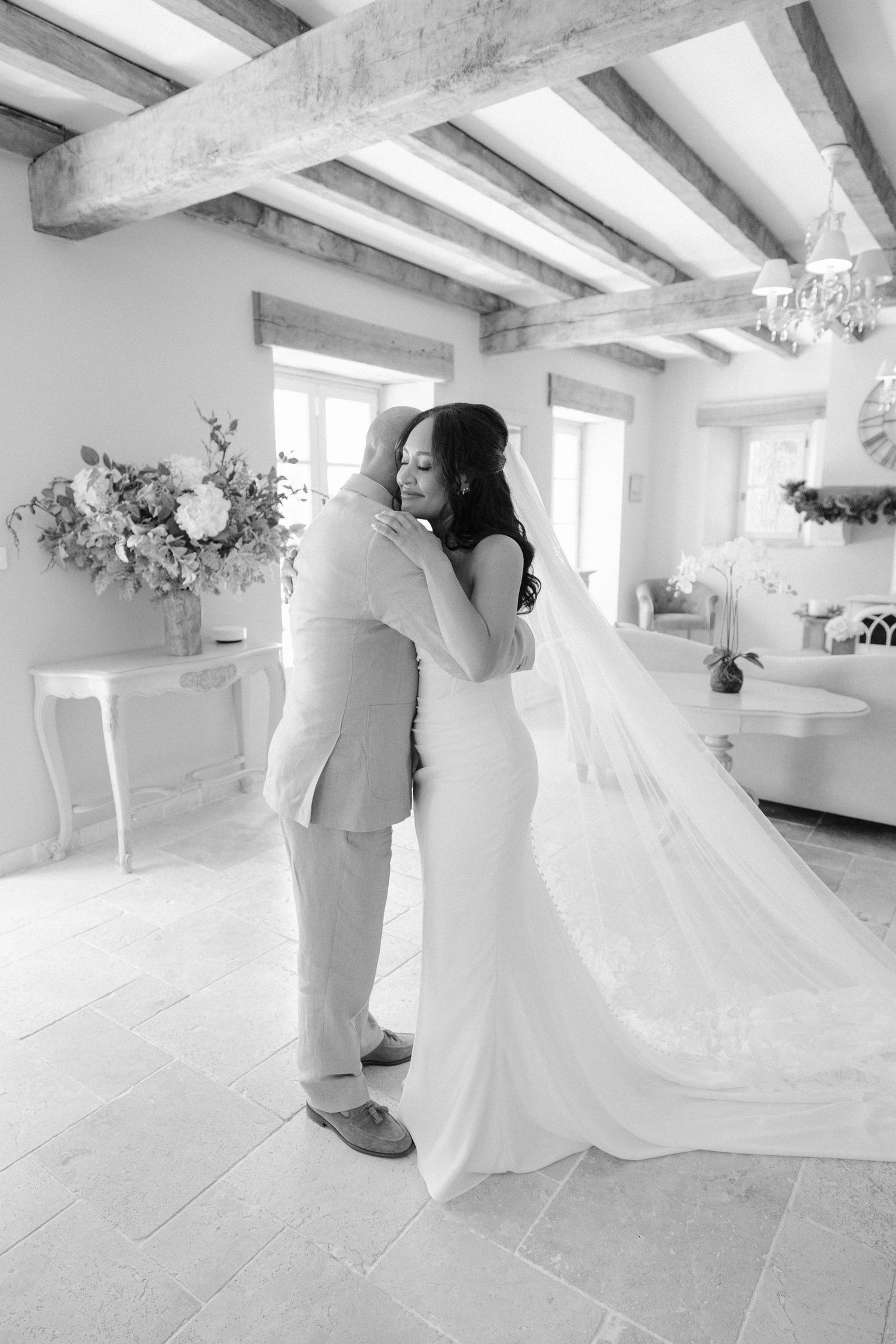Black and white photo of bride in fitted gown and veil sharing tender first look embrace.