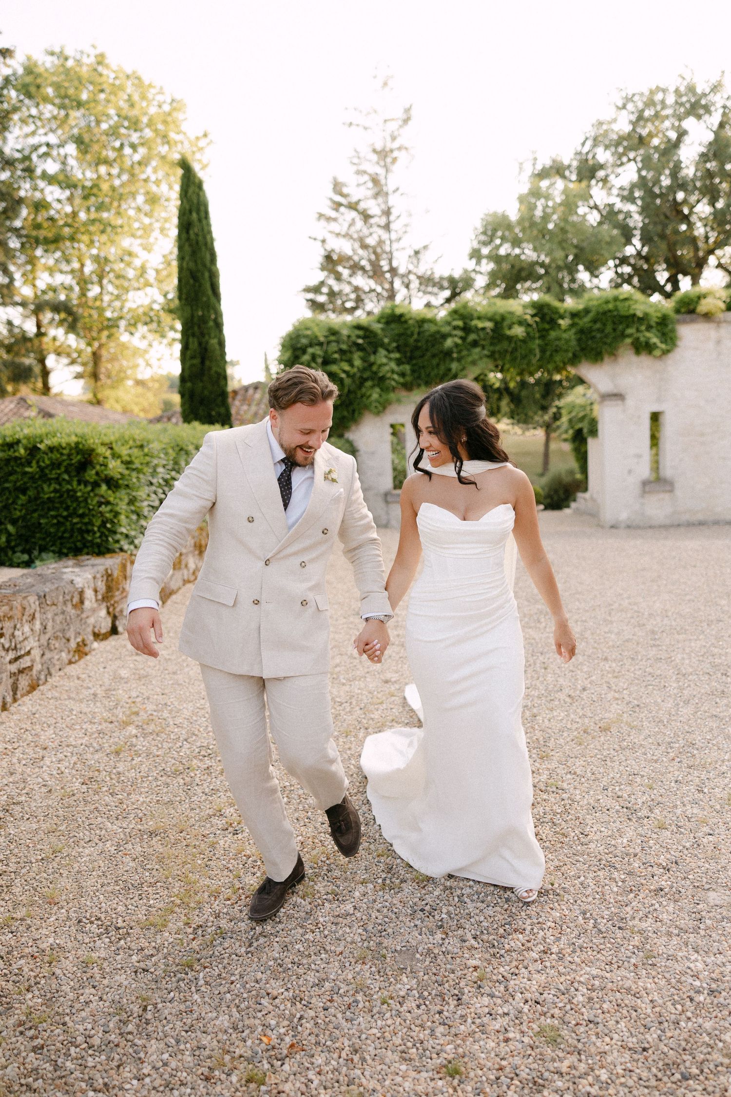 Joyful bride and groom in elegant wedding attire holding hands and walking on a gravel path at a French chateau.
