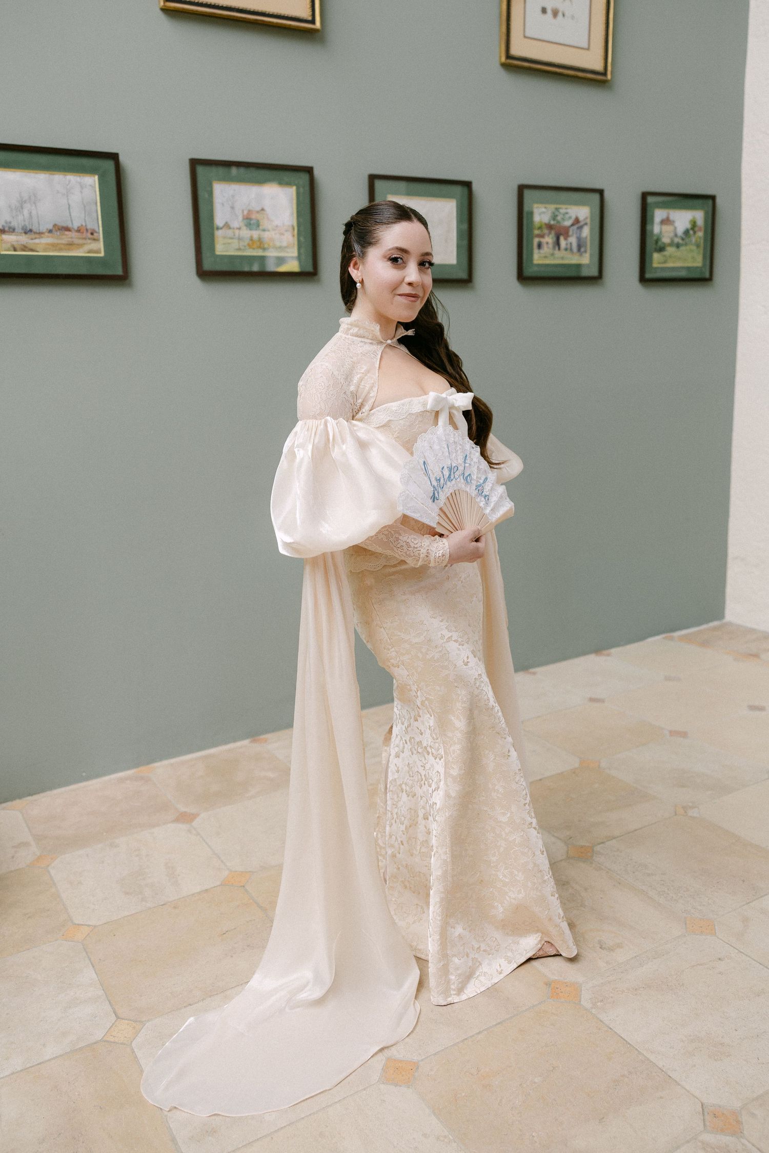 Bride in ivory lace mermaid gown with sheer sleeves holding a blue fan in an art gallery setting.