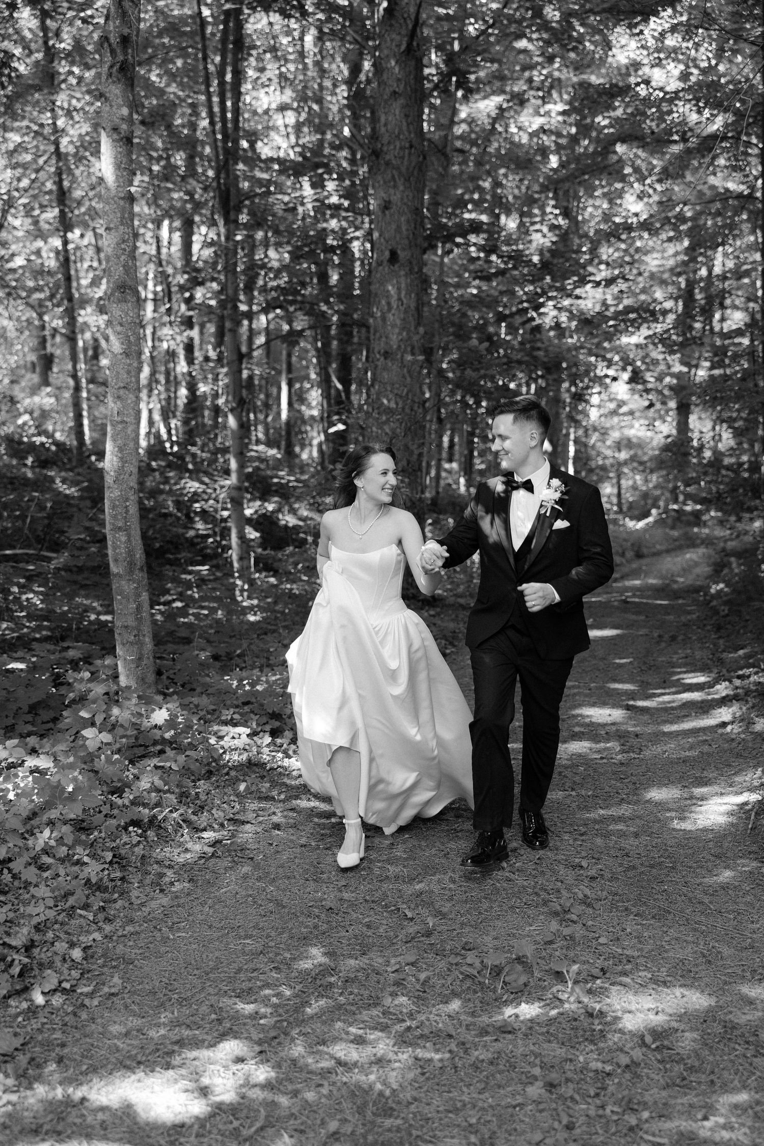 Black and white photo of a smiling bride and groom walking hand in hand along a sunlit forest path.