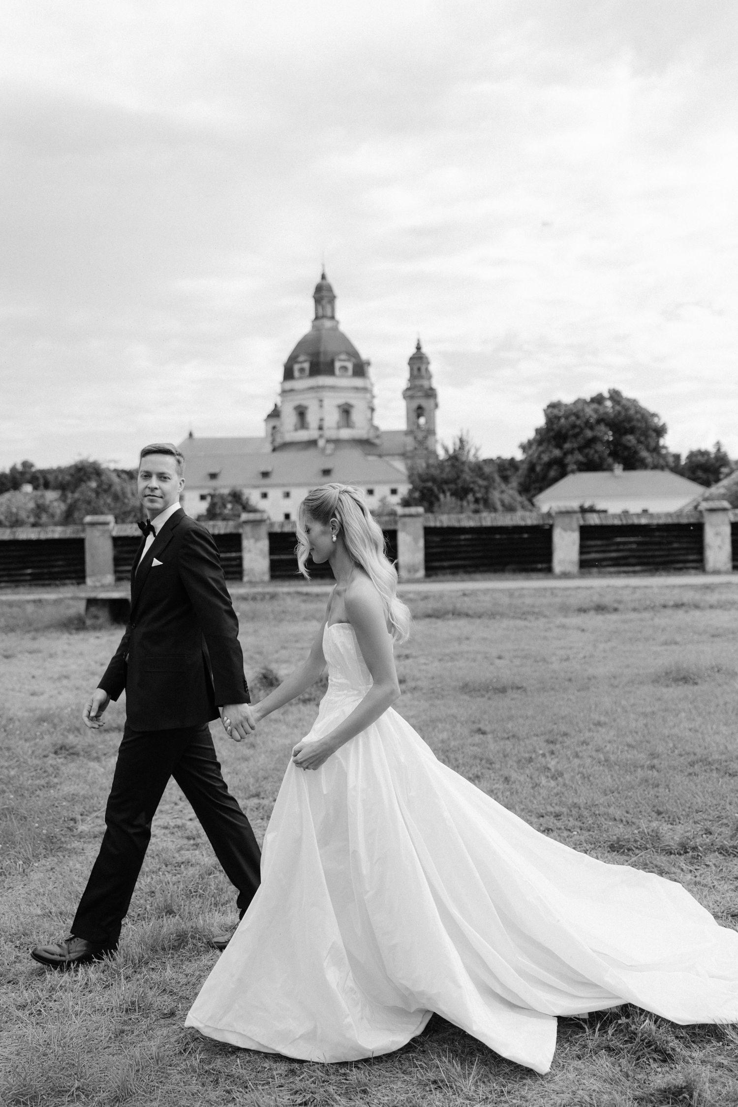 Black and white photo of bride and groom walking hand in hand near a historic church.
