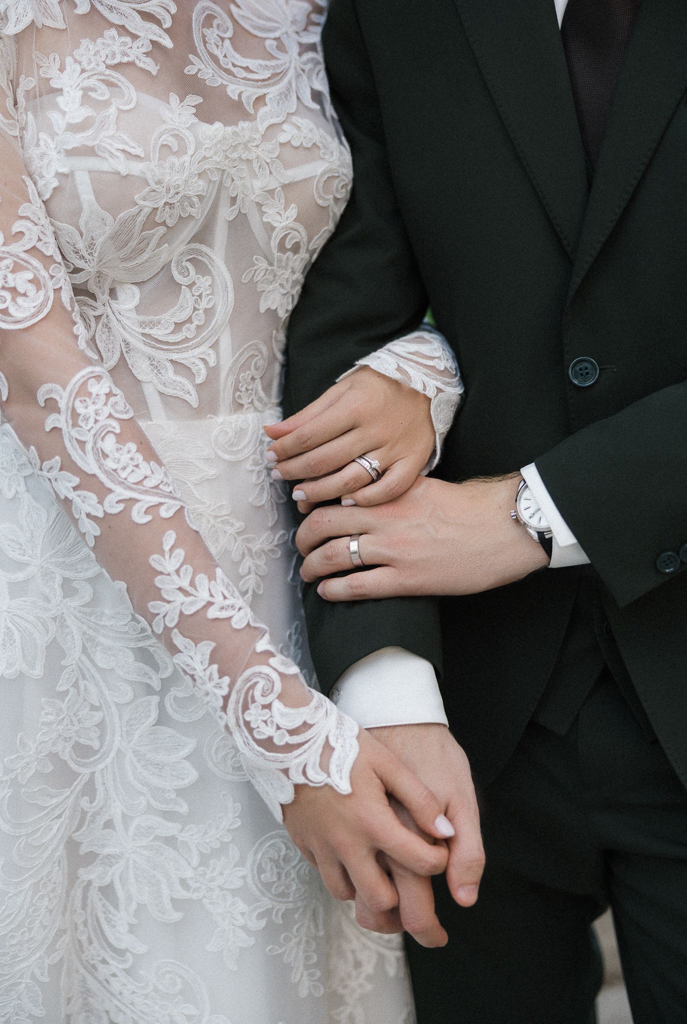 Close-up of newlywed couple's hands with wedding rings, showing intricate lace sleeve detail and dark suit jacket.