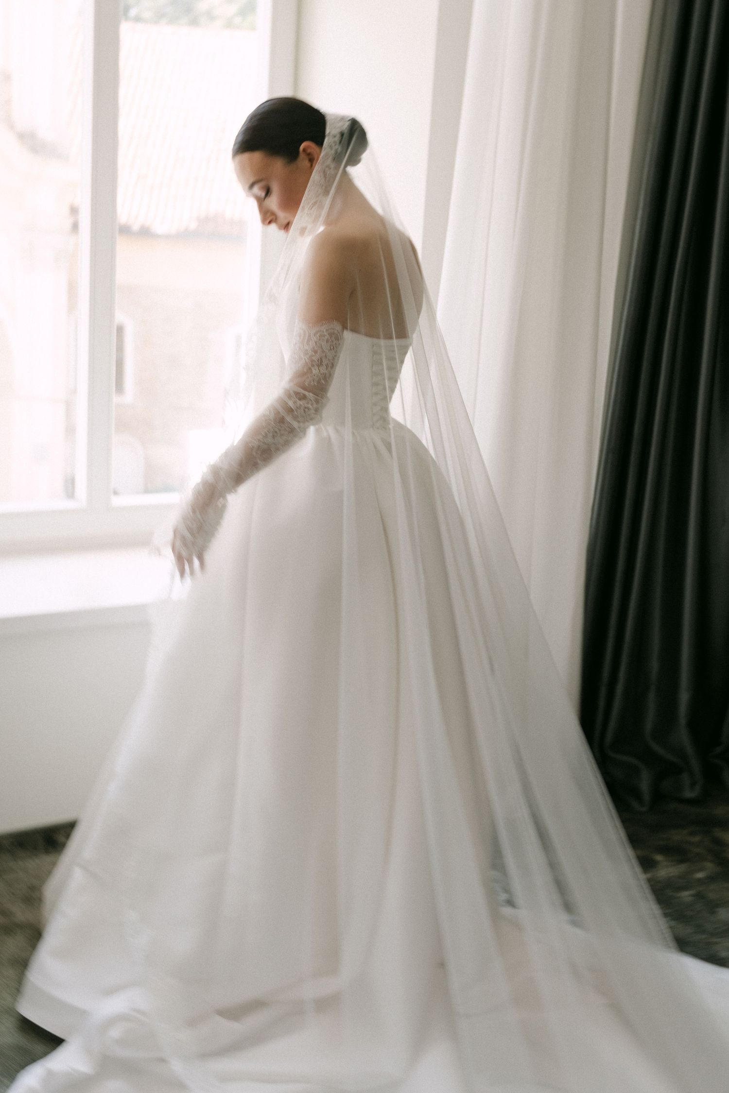 Bride in off-shoulder ball gown with lace gloves and cathedral veil stands by window in elegant bridal suite.