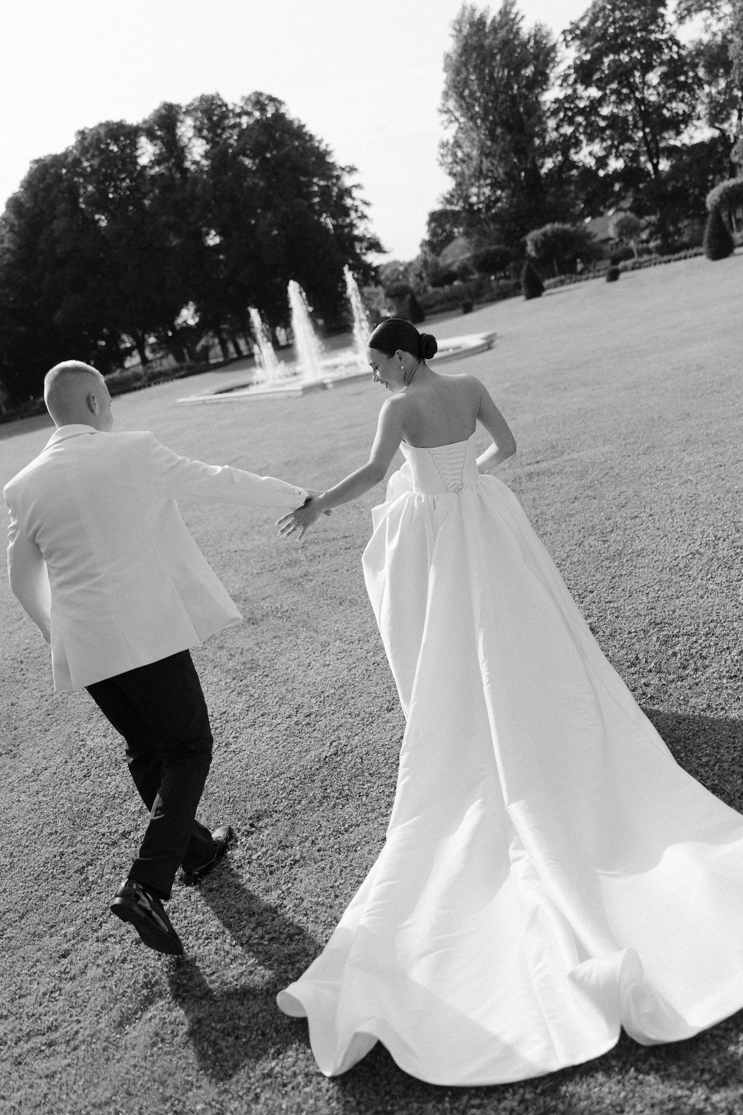 Black and white photo of bride and groom walking hand in hand near a fountain in a park.