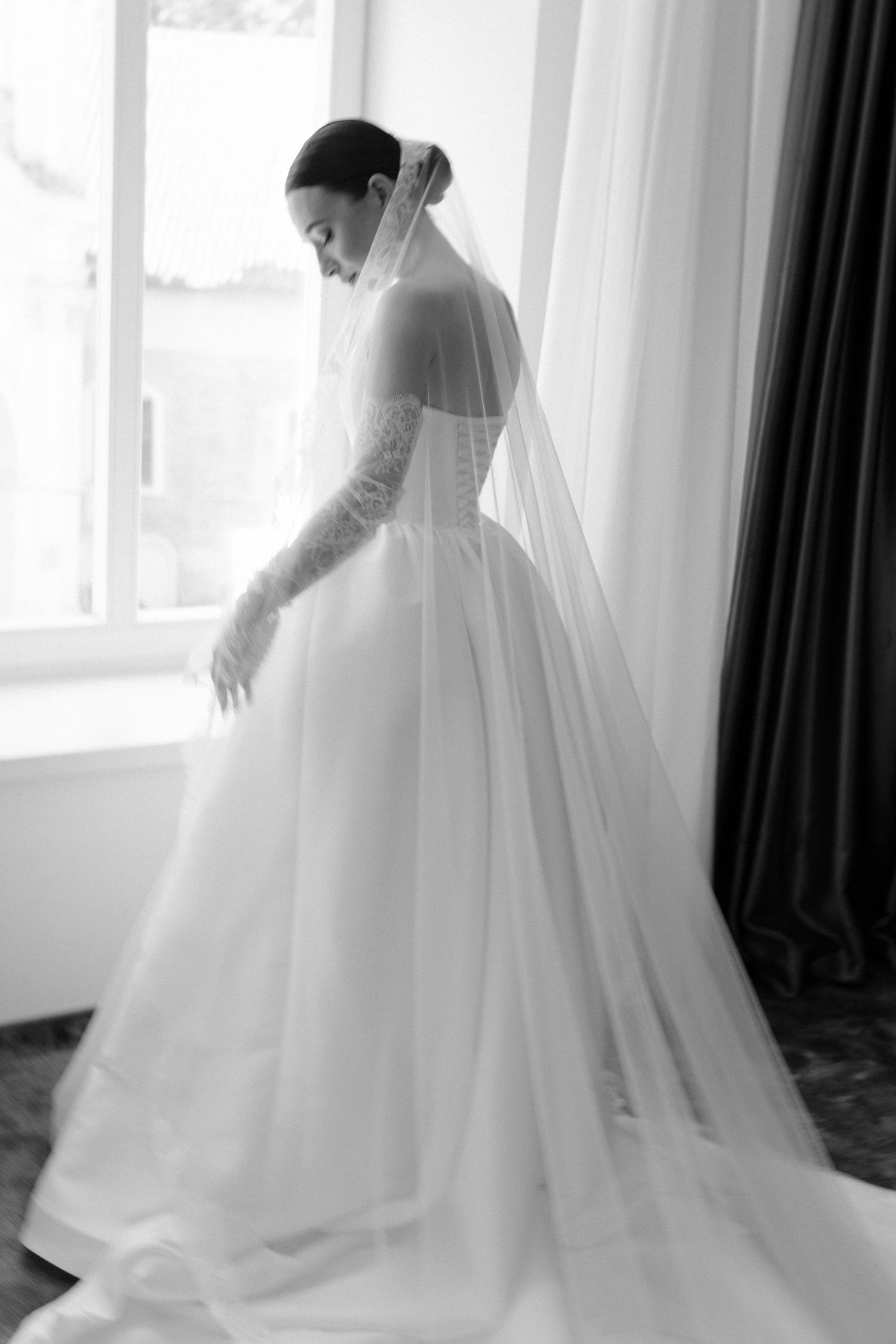 Black and white portrait of a bride in a ballgown with long veil standing by a window.