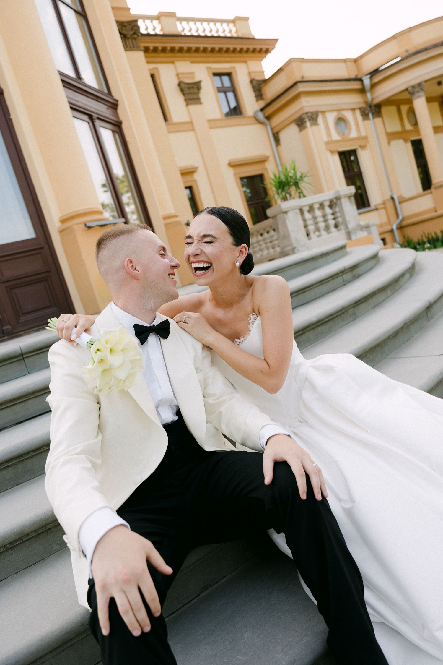 Laughing bride and groom sitting on grand staircase steps outside a yellow neoclassical wedding venue.