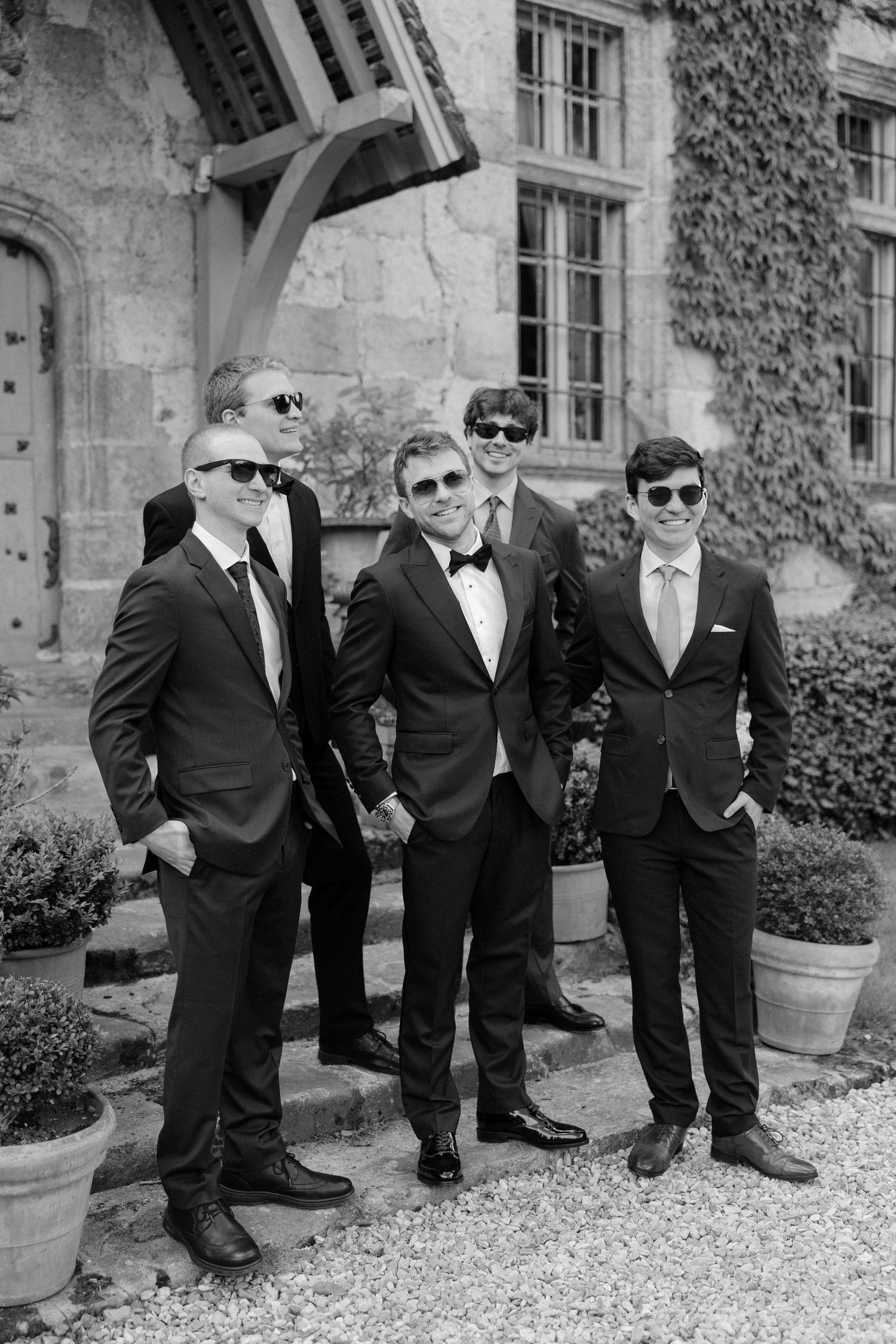 Groom and groomsmen in black suits and sunglasses posing outside a stone manor in black and white.