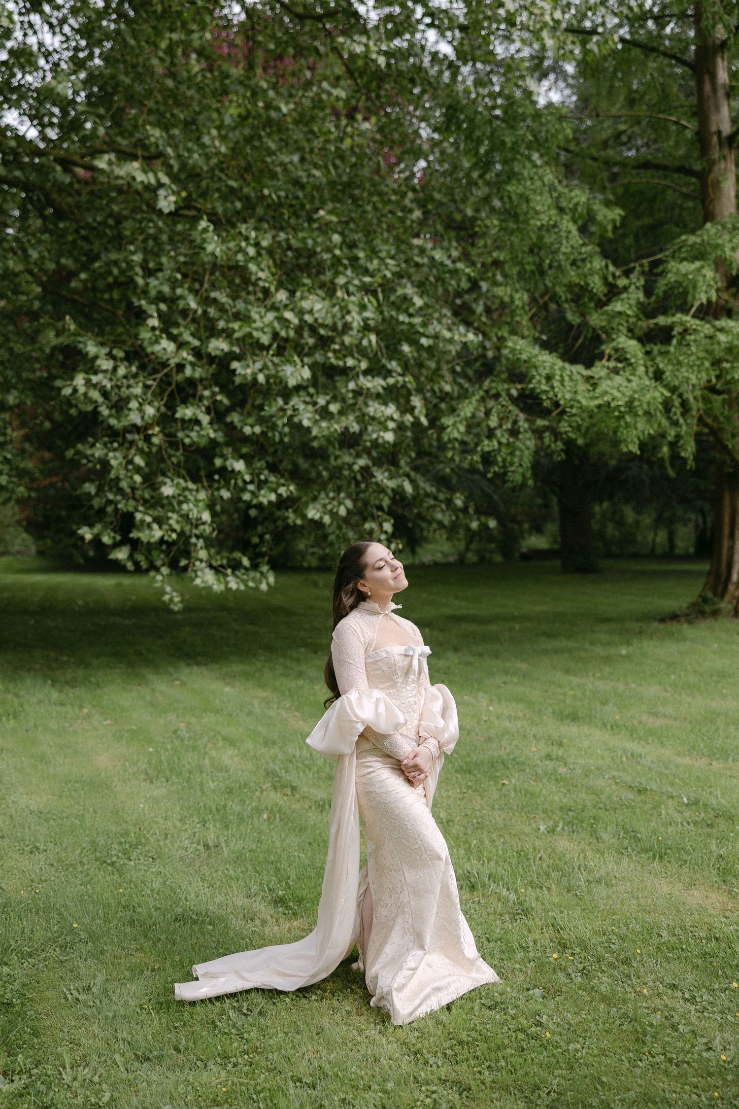 Elegant bride in long-sleeve ivory gown with train stands gracefully on lush green lawn among tall trees.