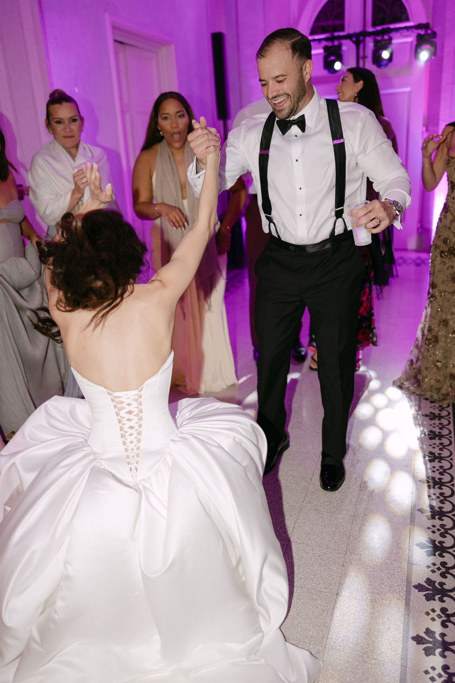 Bride and groom dance energetically on a lit dance floor surrounded by cheering wedding guests.