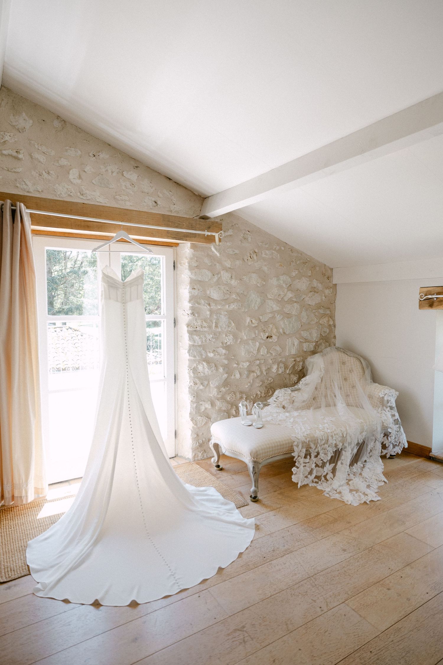 Elegant wedding dress with long train hanging in a rustic stone-walled bridal suite with wooden floors.