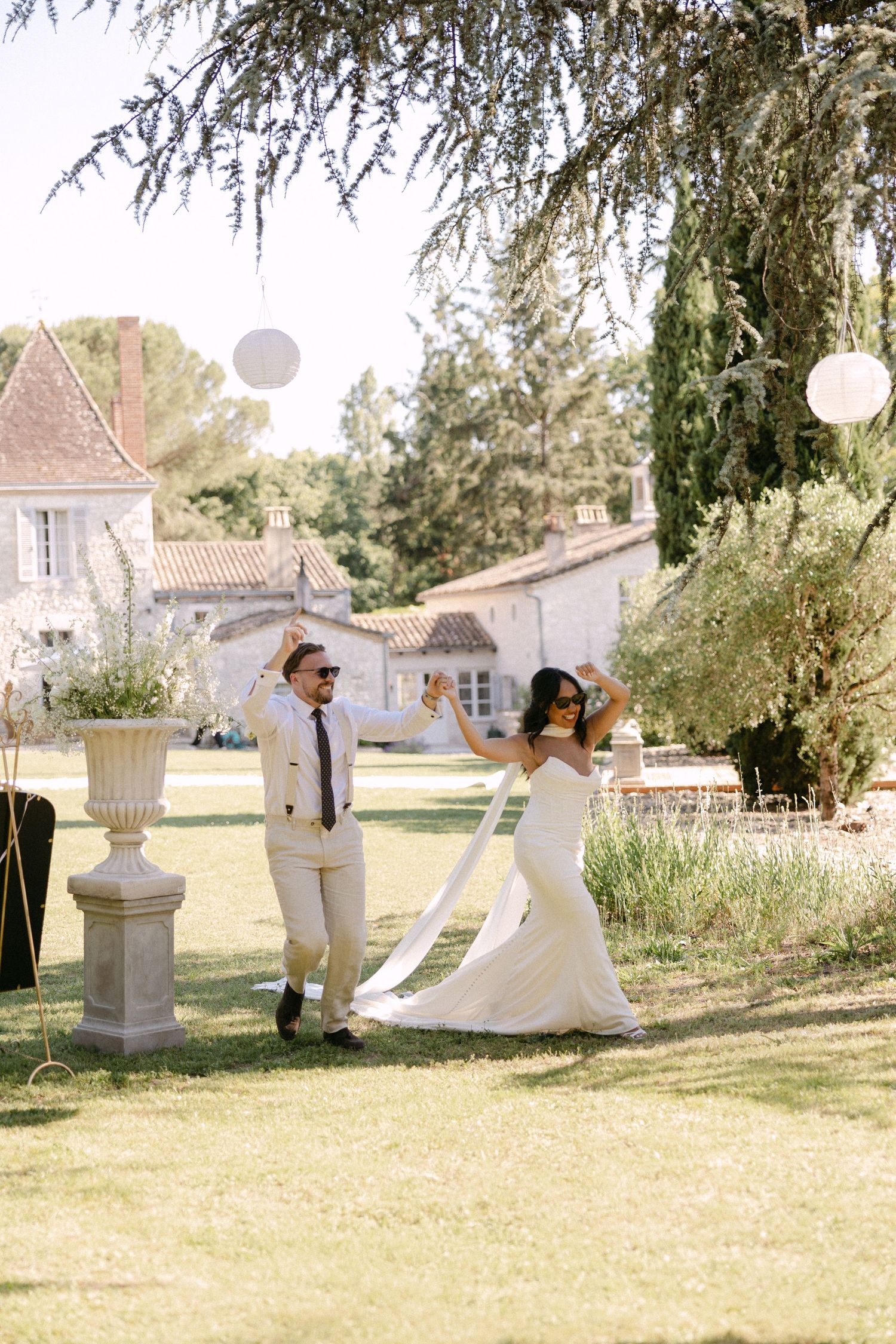 Bride in white gown and groom in beige suit dancing joyfully on a sunlit chateau lawn.