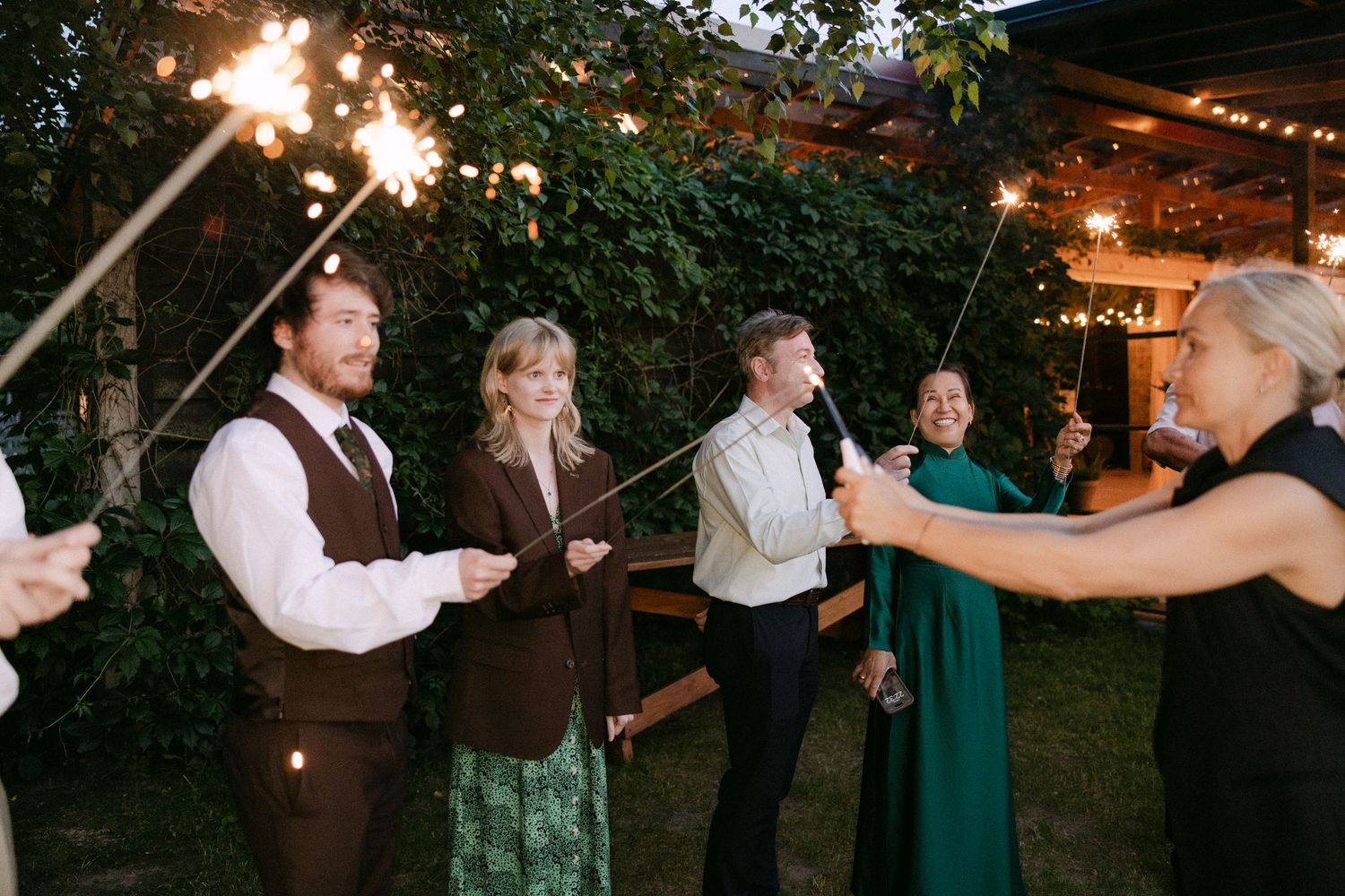 Wedding guests holding sparklers celebrate outdoors at evening reception surrounded by string lights.