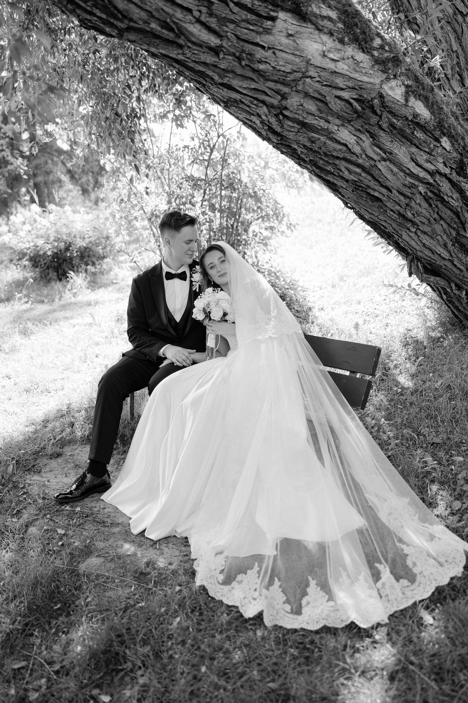 Black and white photo of bride and groom sitting romantically on a bench beneath a large tree in nature.