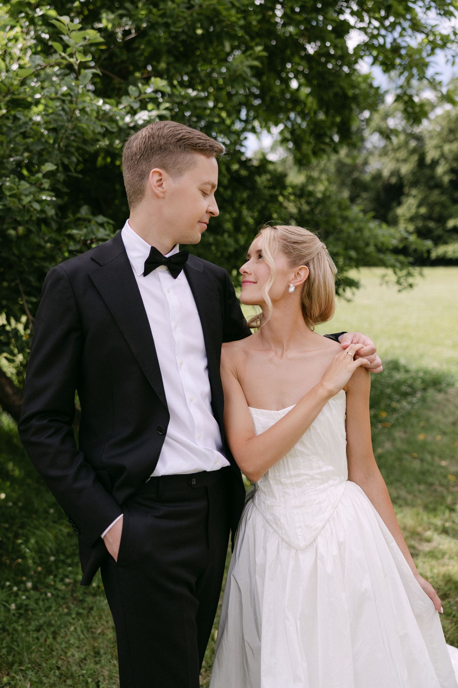 Elegant bride and groom share a tender moment outdoors, she in strapless white gown, he in classic black tuxedo with bow tie.