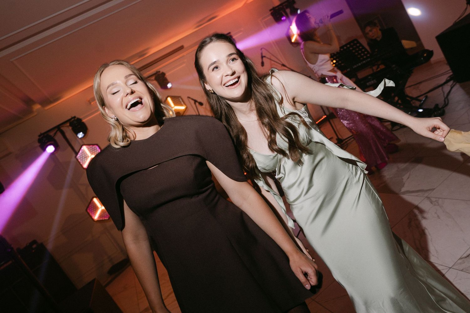 Two joyful women in elegant dresses laughing and dancing together at a wedding reception party.