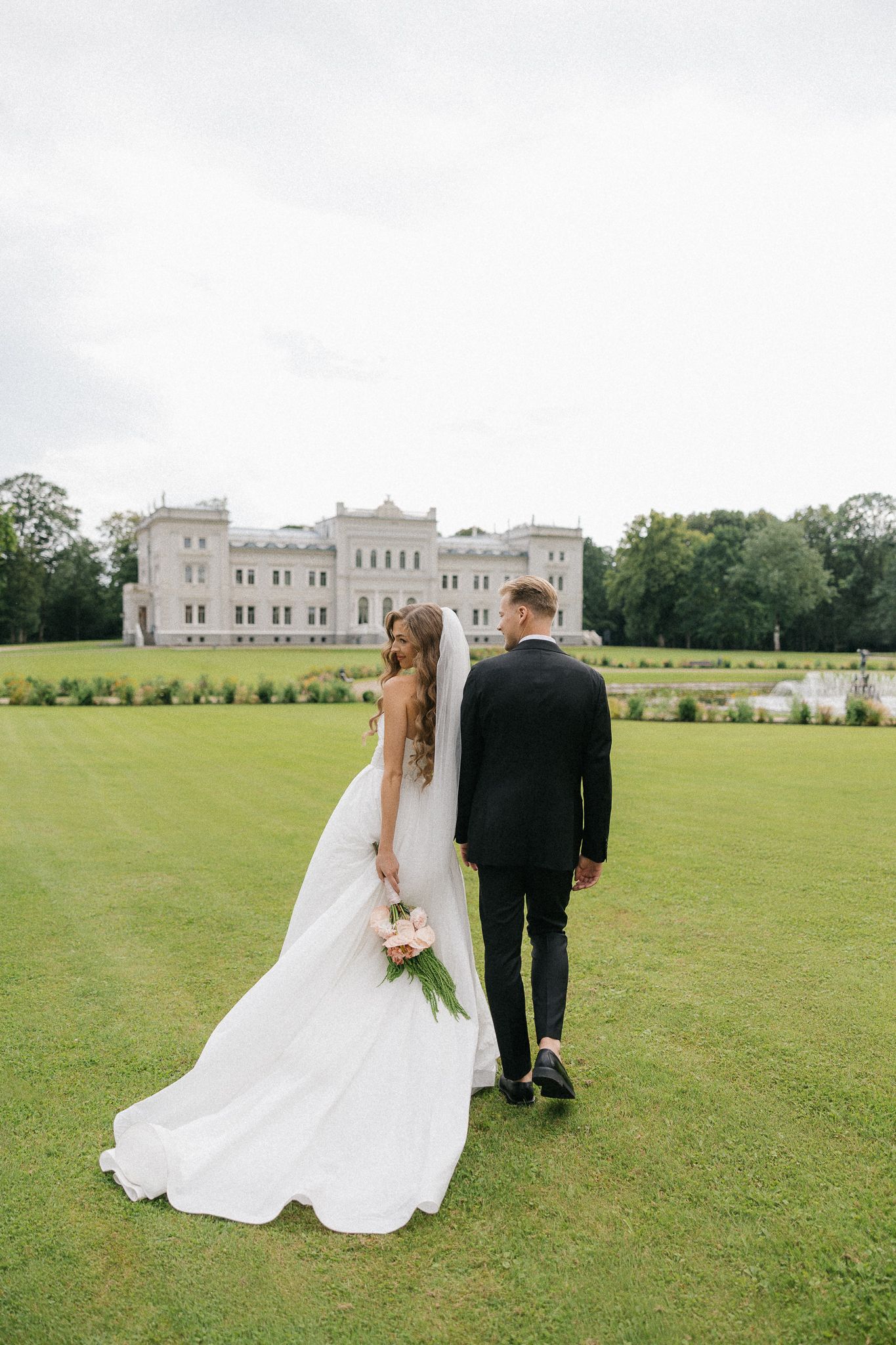 A wedding couple stands on manicured lawn in front of a grand white castle-like mansion on their special day.