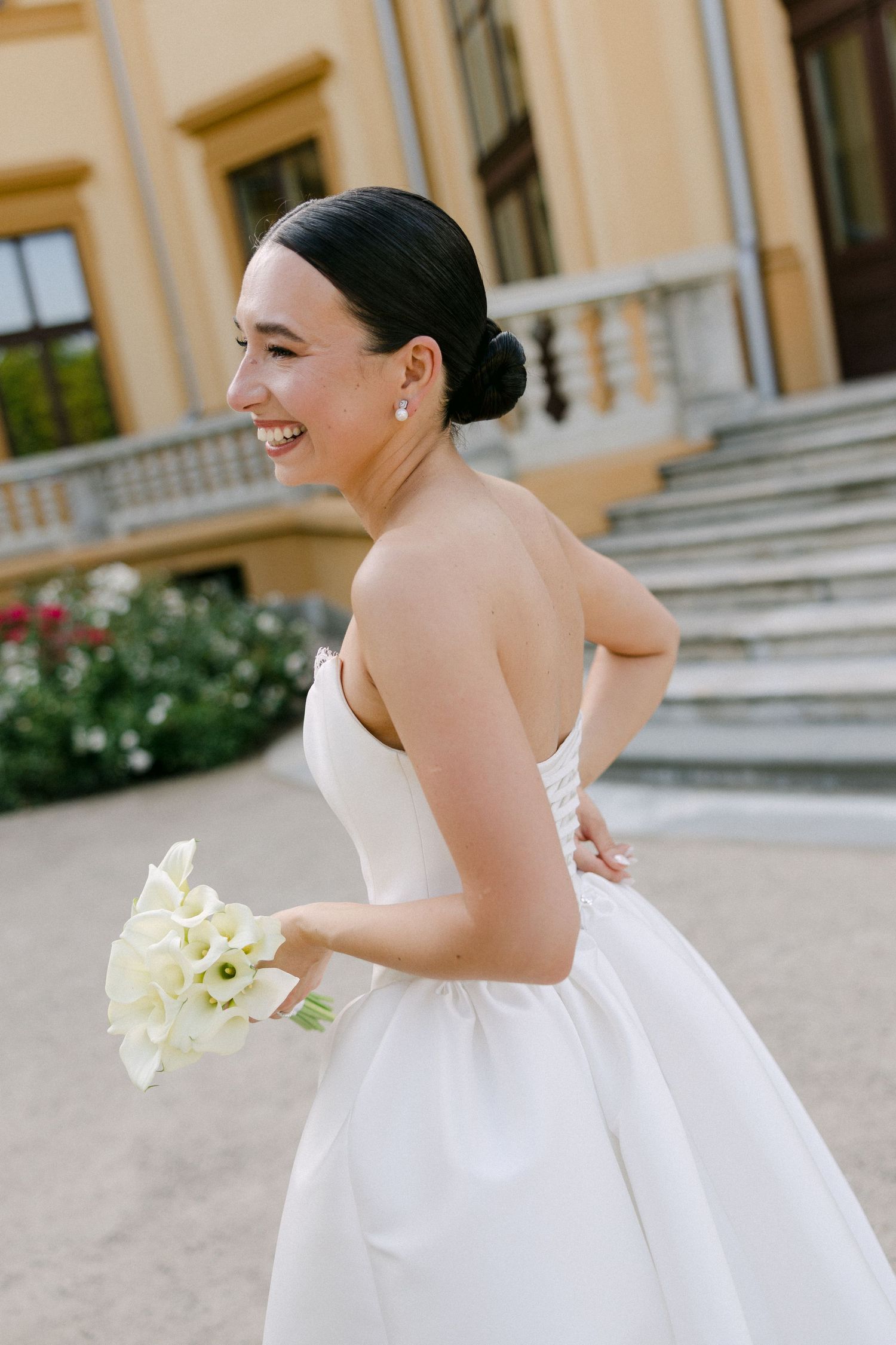 Smiling bride in a strapless white gown holds white calla lily bouquet outside a grand yellow building.