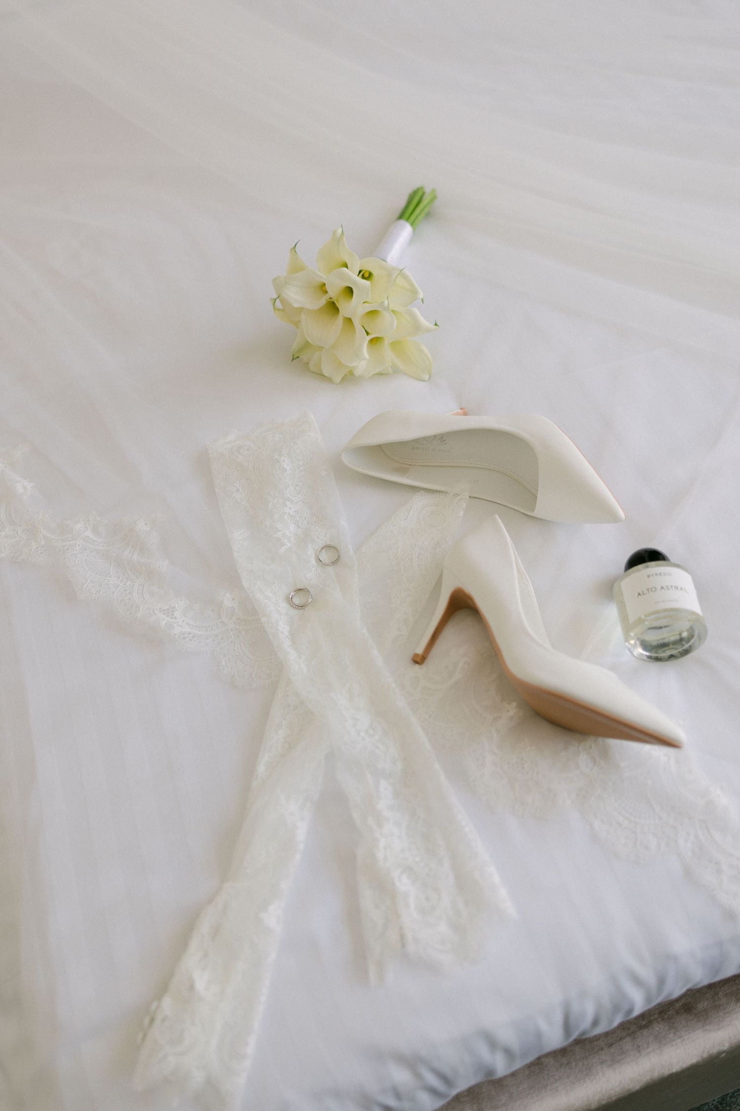 Elegant wedding flat lay with white heels, lace veil, calla lily bouquet, rings, and perfume on a bed.