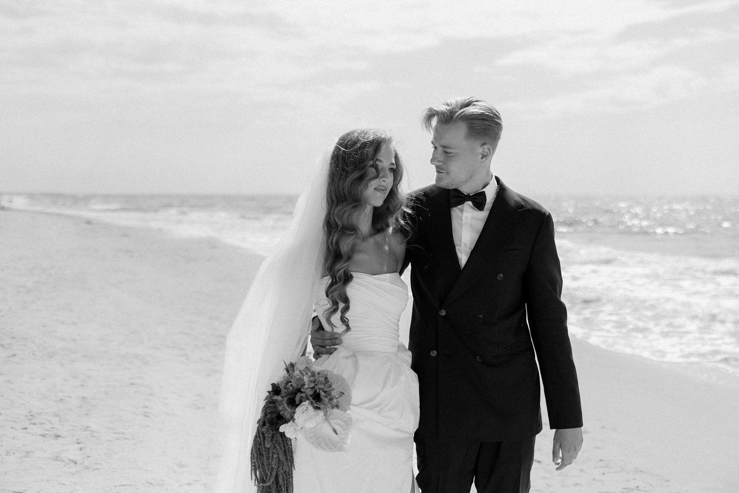 A couple in formal wedding attire walks along a misty beach shoreline at sunset.