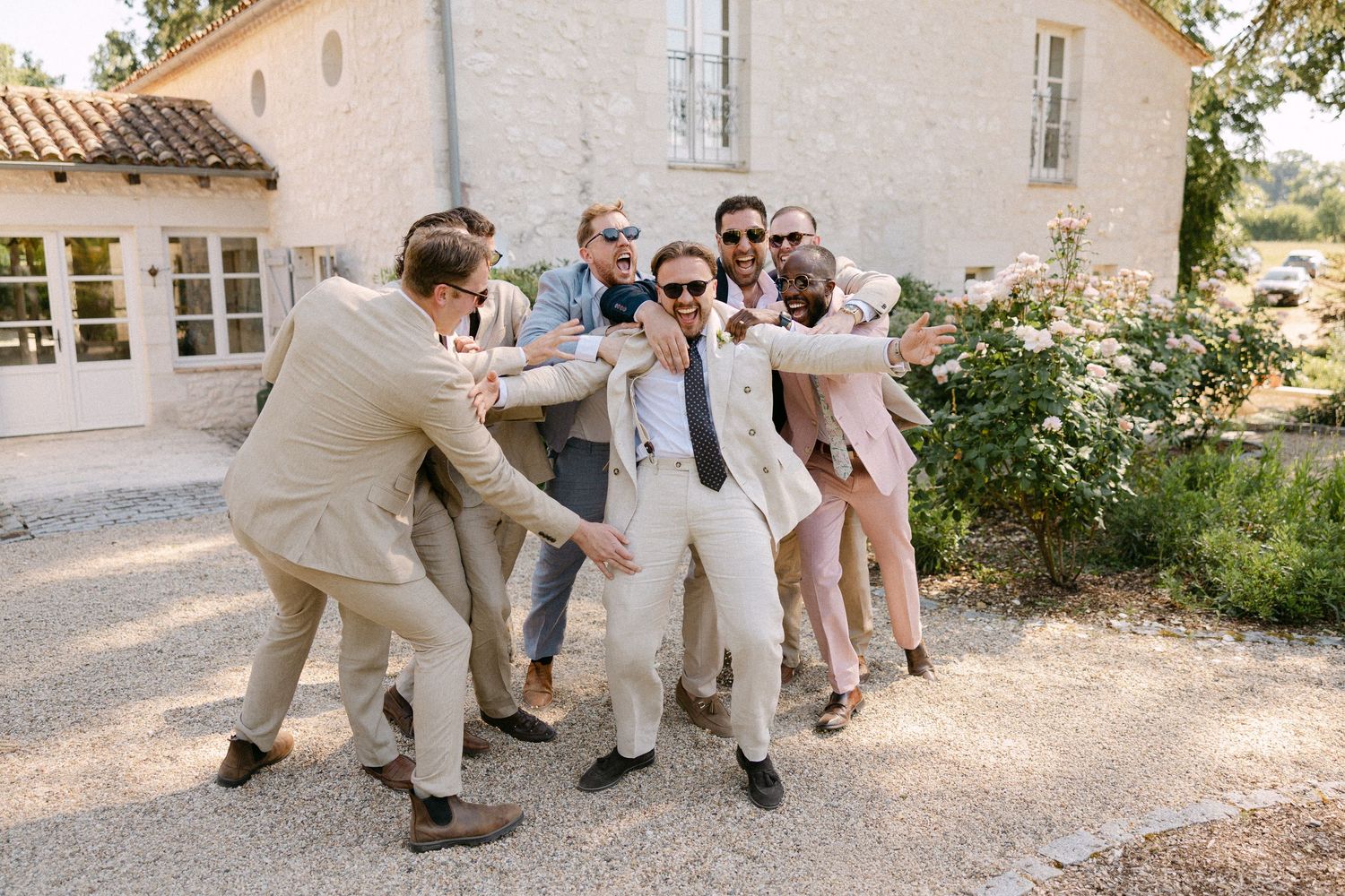 Groom in white suit surrounded by laughing groomsmen in beige suits outside a French estate.