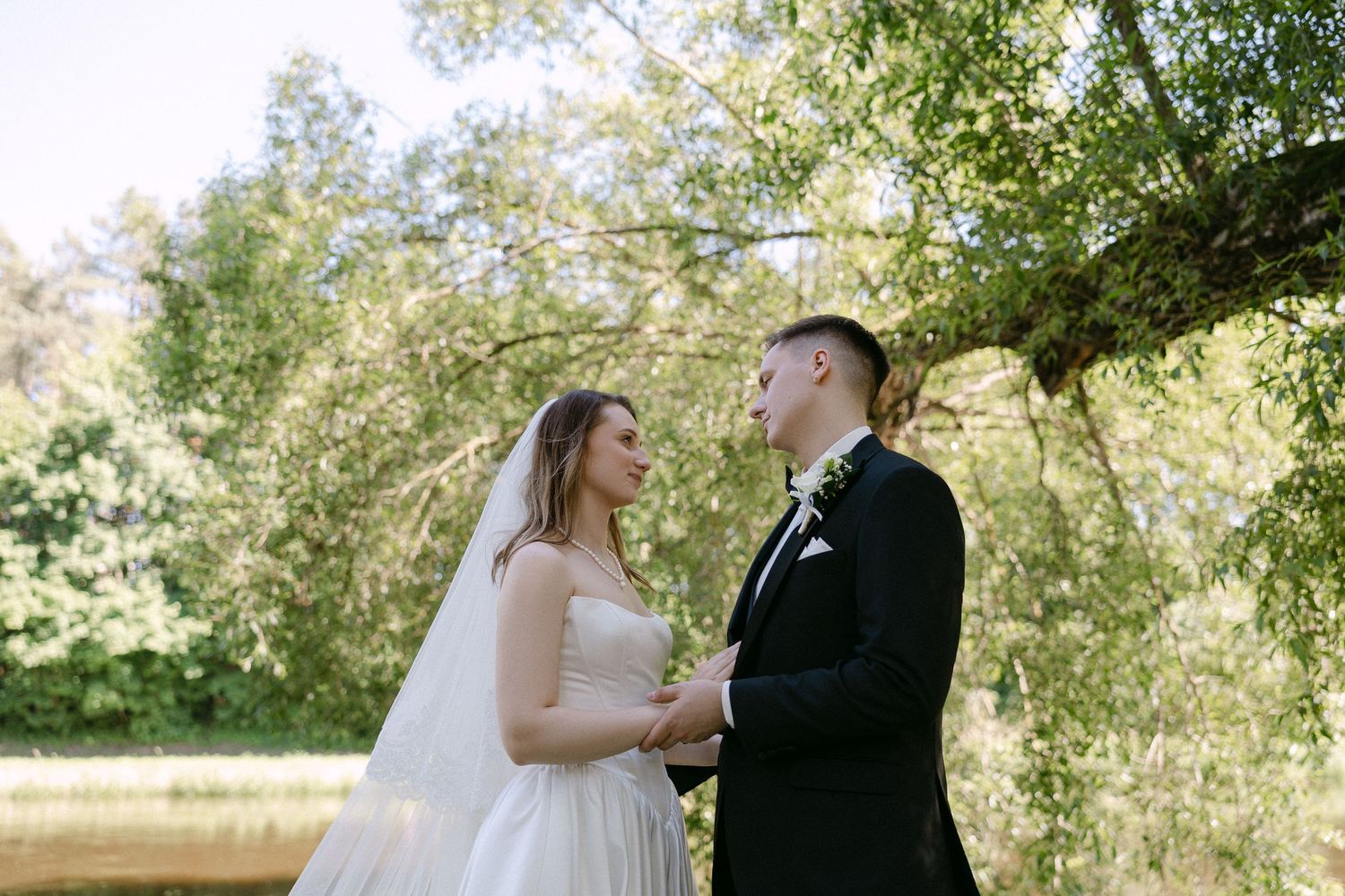 Bride and groom holding hands gazing at each other under lush green trees by lake.
