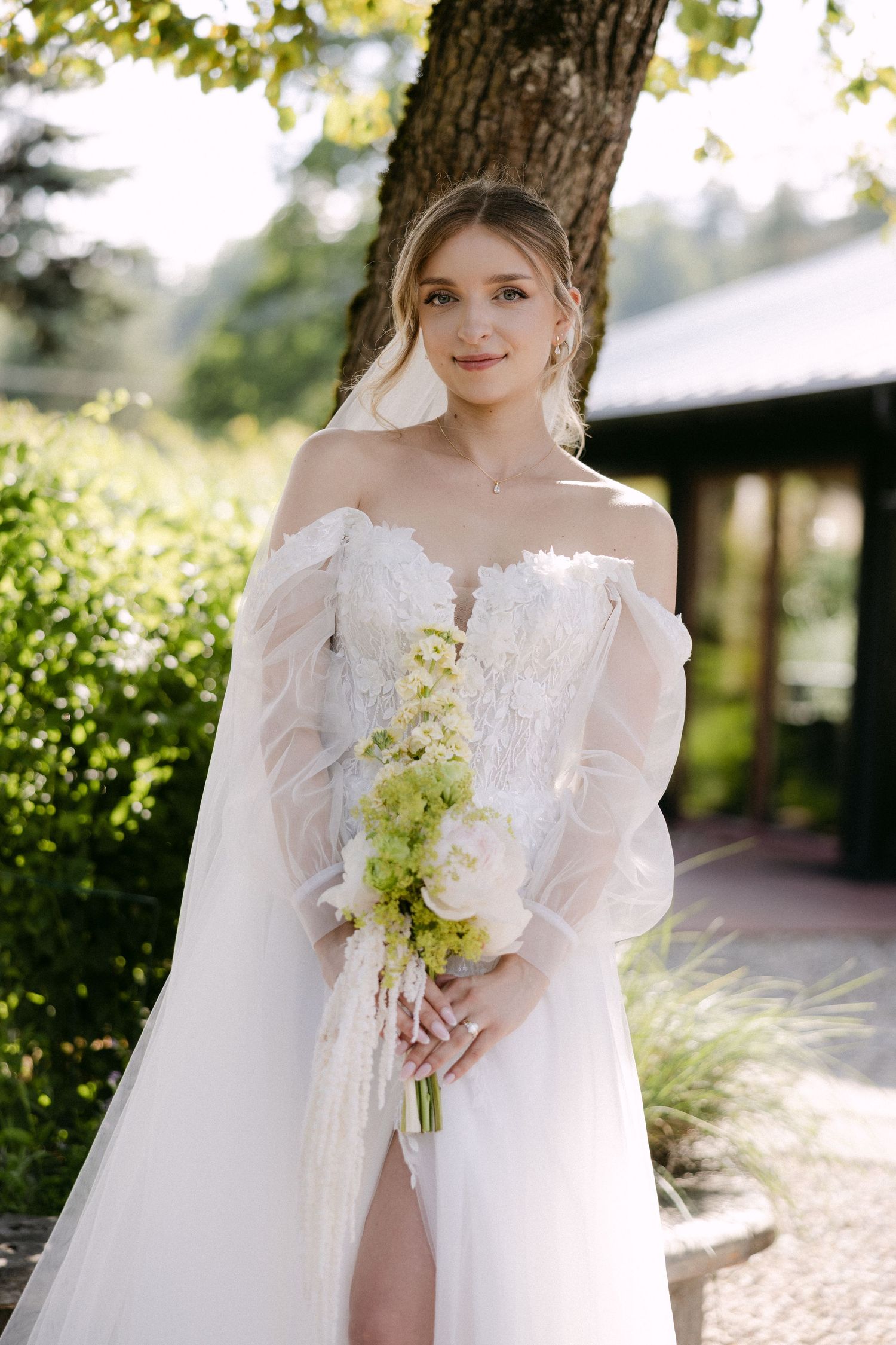 Bride in off-shoulder white lace gown holds green and white bouquet outdoors near lush garden trees.