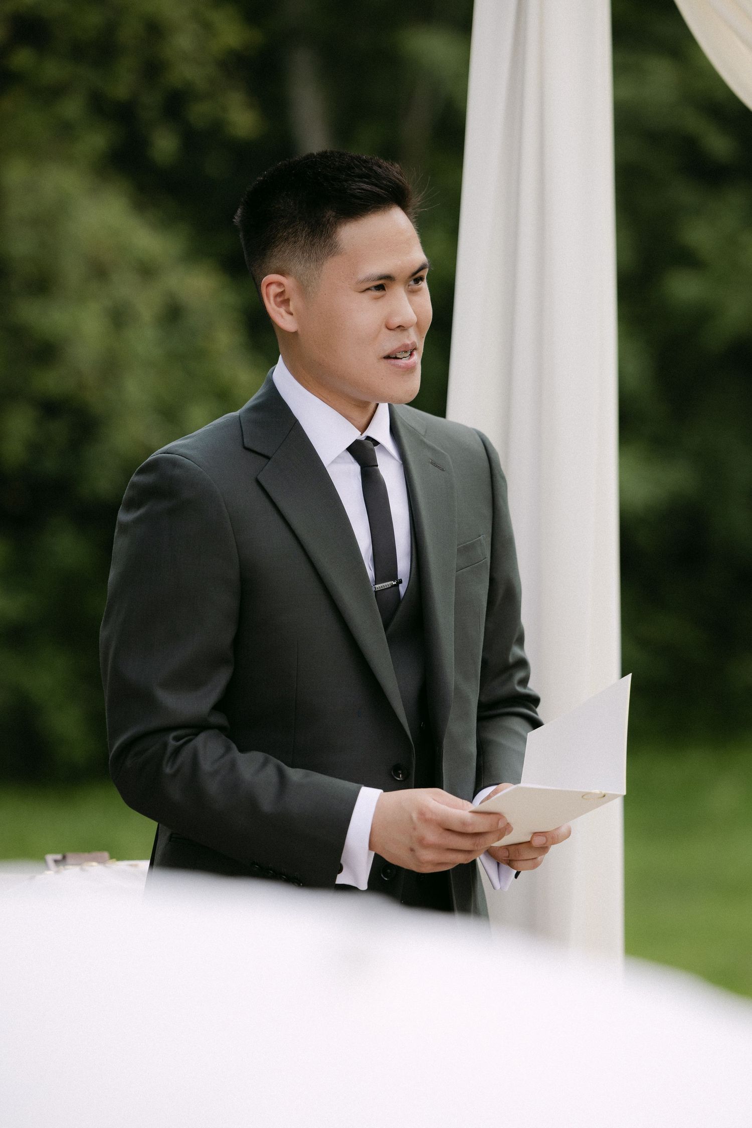 Asian groom in charcoal suit reading vows at outdoor wedding ceremony beside white floral arch.