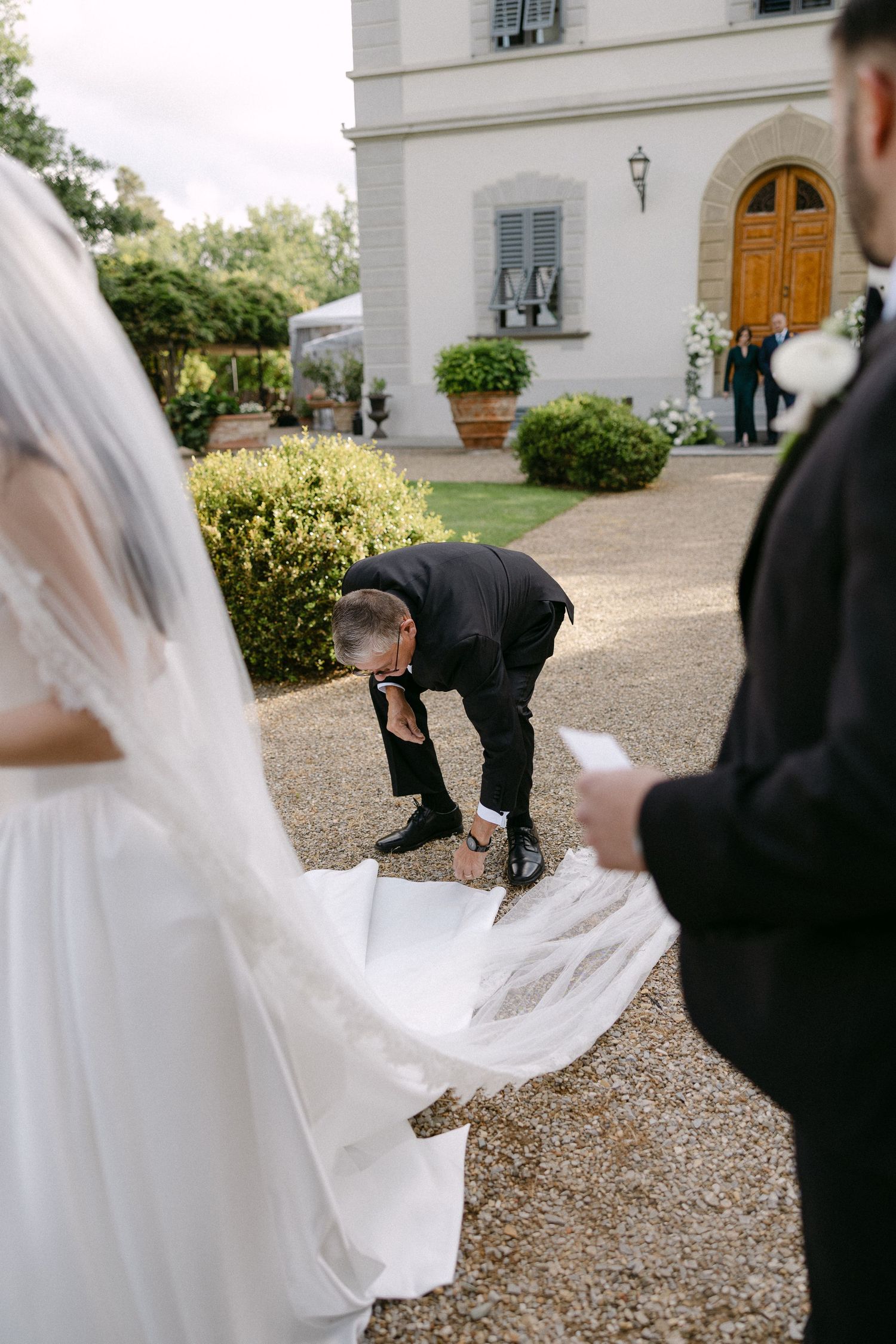 Wedding attendant adjusts bride's white gown train on gravel path outside elegant villa.